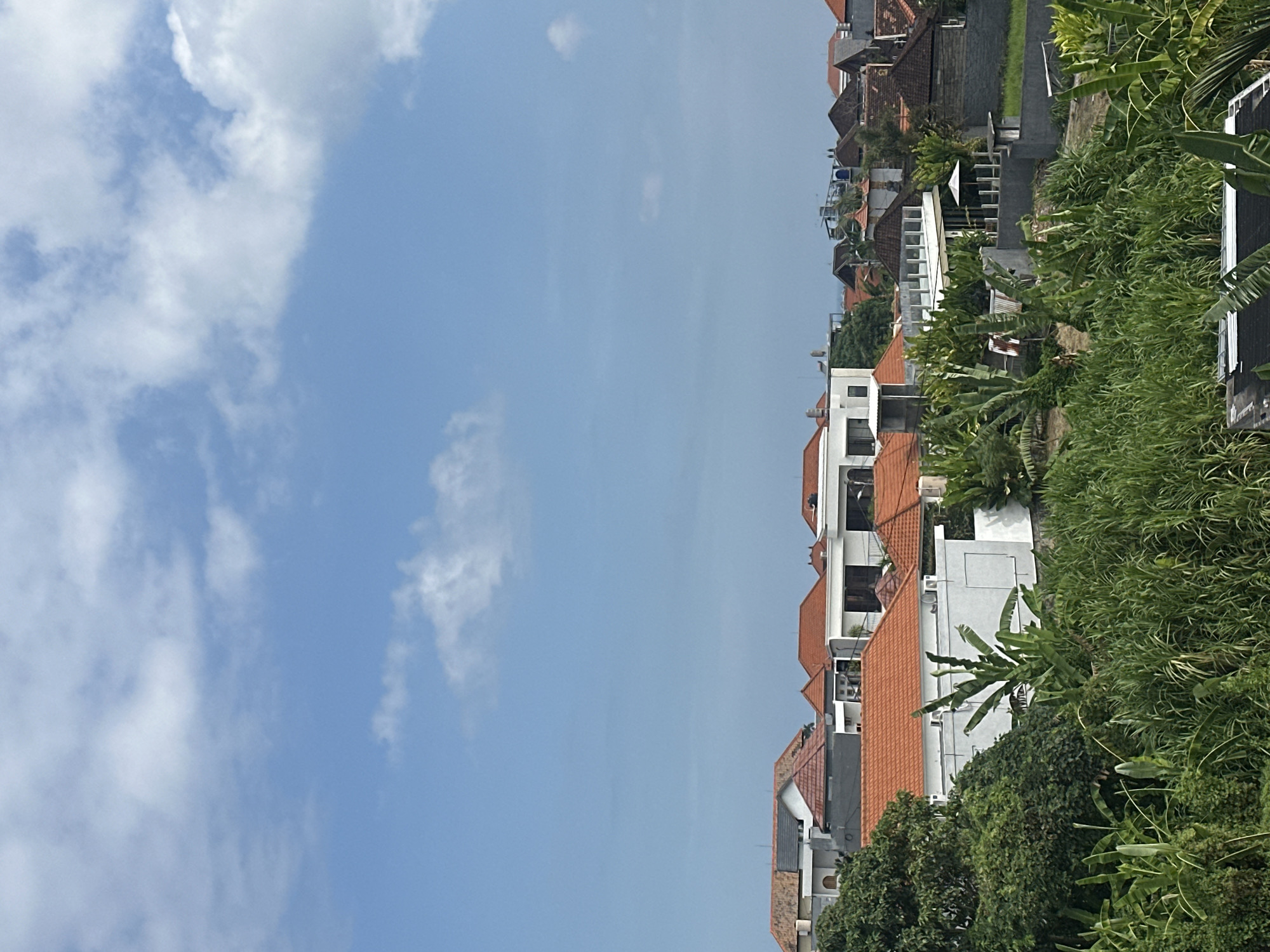 The image shows a view of North Kuta, Indonesia. The sky takes up a large portion of the scene, with white clouds scattered across a blue background. On the right side, there are closely packed buildings with orange tiled roofs and white walls, mixed with lush tropical vegetation. The buildings appear to be multi-story, some with visible balconies. The vegetation includes trees and plants with large, broad leaves, creating a dense, green barrier along the edge of the frame.
