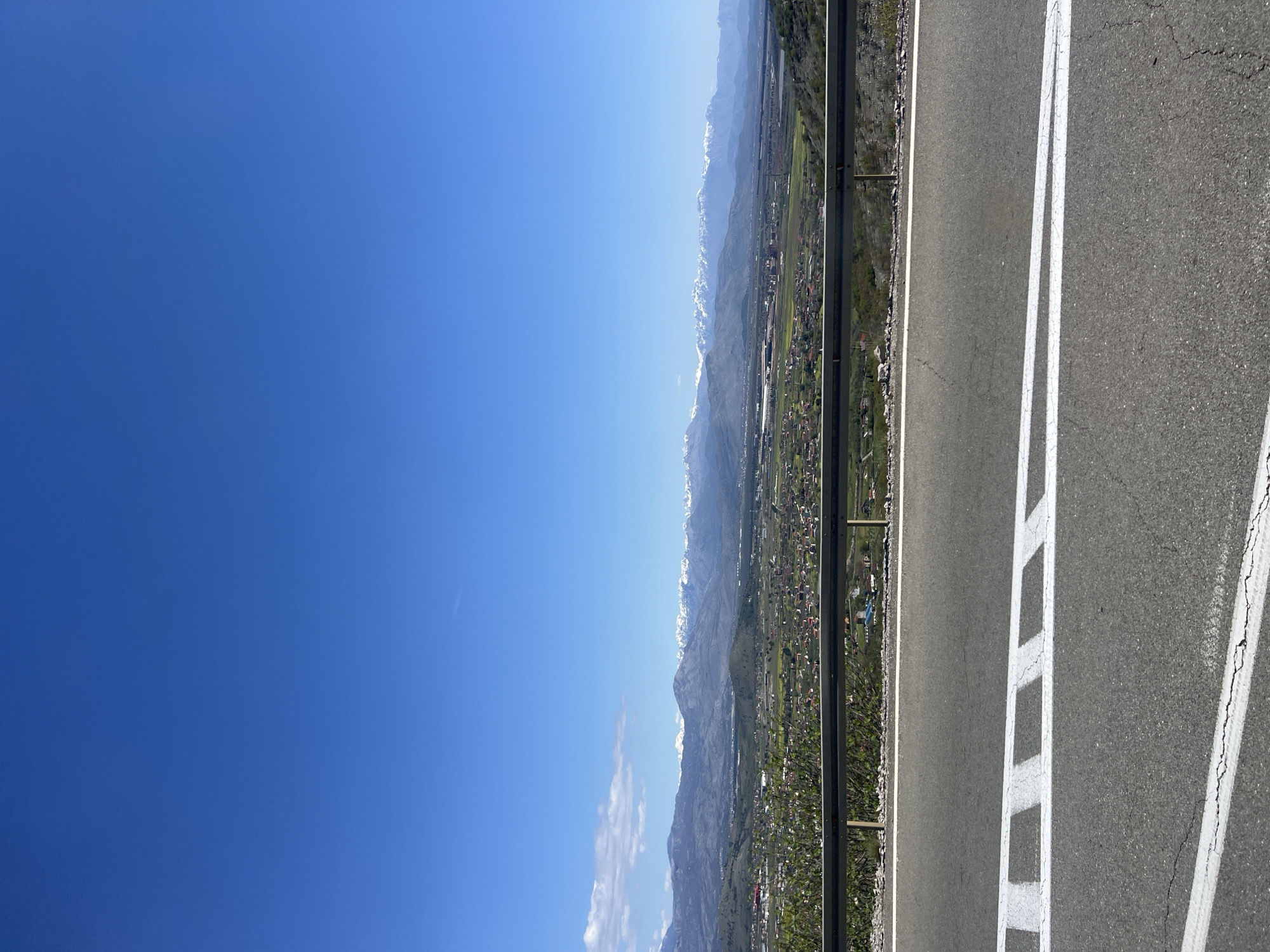 The image shows a view from a road in Gornji Kokoti, Montenegro, overlooking a valley with a town and mountains in the background. The sky is clear and blue. The road is paved with asphalt and has white lane markings. A metal guardrail runs along the edge of the road. The town in the valley appears to have houses with red roofs and patches of greenery. The mountains in the background are partially covered with snow.