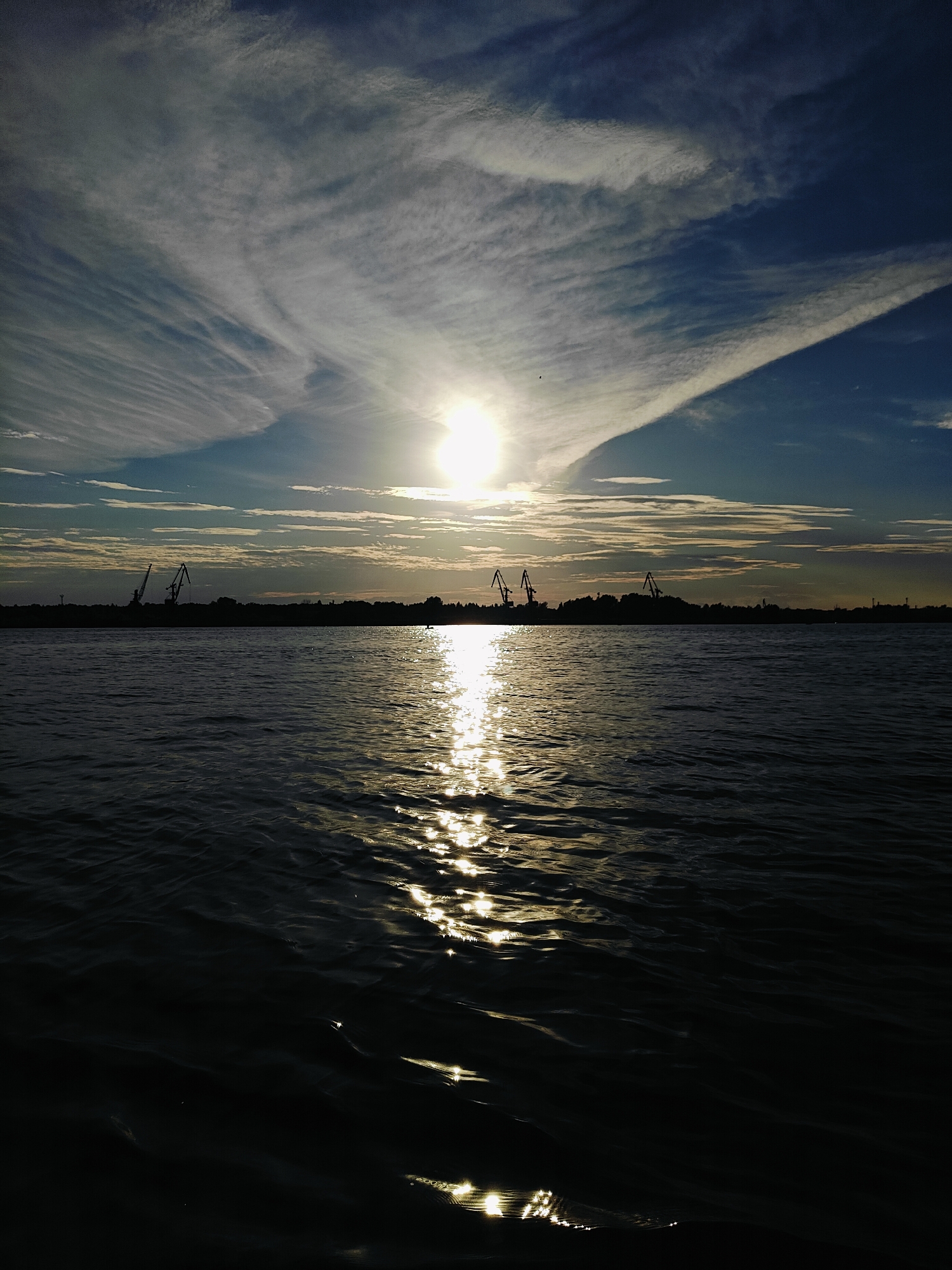 The image depicts a sunset over a body of water, likely a river or reservoir, with the silhouette of a coastline in the background. The sun, partially obscured by wispy clouds, is low on the horizon, casting a bright reflection on the water. The sky is a gradient of blue and white, with streaks of clouds that appear to be fanning out from the sun.
Along the distant coastline, several cranes are visible, suggesting a port or industrial area. The silhouette of trees and other structures can also be discerned against the twilight sky. The water itself is dark, with ripples and glints of light from the setting sun.
The scene is likely located along the Kama River, which flows through Chaikovsky, Russia. The presence of cranes indicates a port facility, which could be part of the industrial infrastructure of the city. The overall impression is one of industrial activity set against the natural beauty of a sunset. There are no people or animals clearly visible in the frame. The time of day is dusk. The weather appears clear with some cloud cover. The composition of the image is horizontal, with the horizon line dividing the sky from the water. The focus is on the interplay of light and shadow, and the vastness of the sky and water.