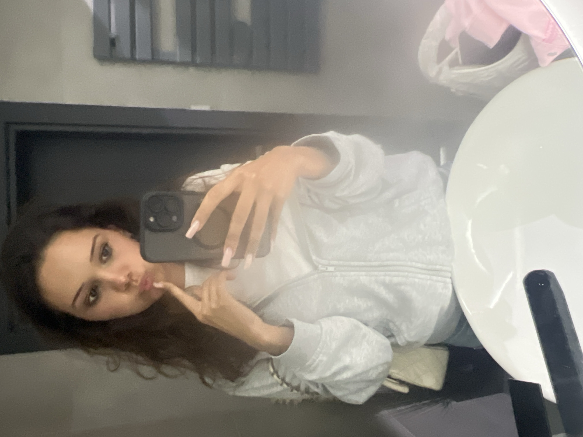 The image depicts a young woman taking a mirror selfie in what appears to be a bathroom. She is wearing a white zip-up hoodie over a white t-shirt and blue jeans. She has long, dark, curly hair and is holding her right index finger to her lips, giving a playful expression. Her left hand is holding a smartphone, which is capturing the reflection. A light-colored quilted bag is slung over her left shoulder. In the background, a dark door frame is visible, and to the right, a radiator and some white bags are present. The foreground shows the edge of a white sink. The image was taken in Zlatograd, Bulgaria.