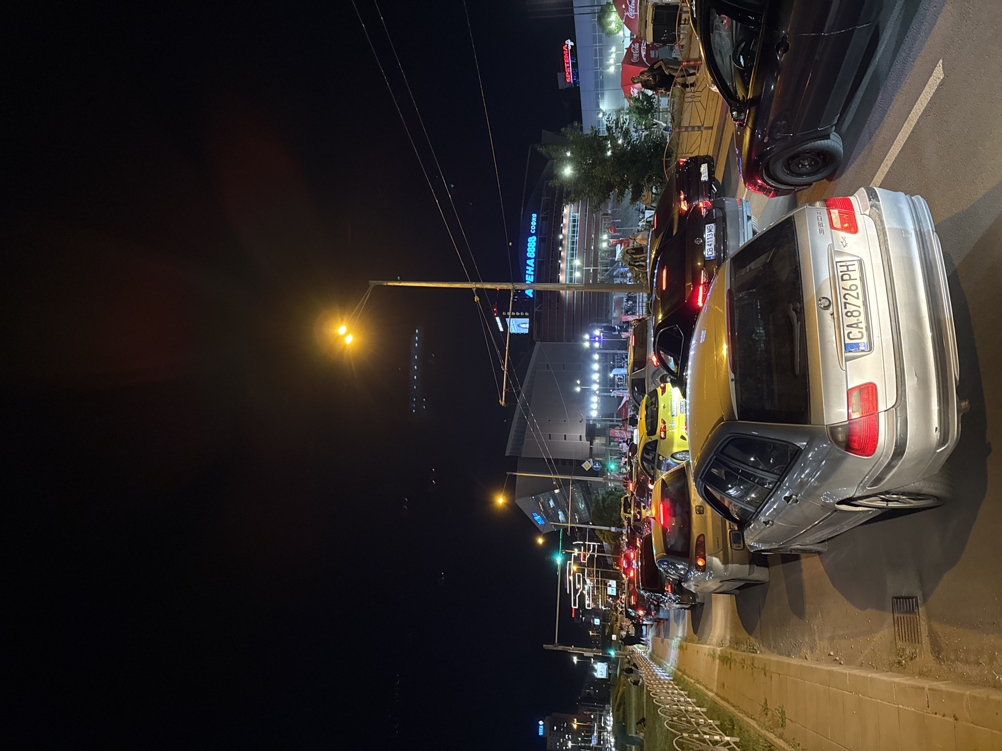 The image depicts a nighttime street scene in Sofia, Bulgaria, characterized by heavy traffic congestion. Multiple vehicles are queued in a line, their tail lights illuminating the scene. The license plate of the silver car closest to the viewer reads "CA 8726 PH" with the Bulgarian flag visible. A building displaying the text "ARENA СОФИЯ" in blue neon is visible in the background. Adjacent to it, the text "SPETEMA" is displayed in red. The environment includes streetlights and other urban infrastructure. People are present on the sidewalks, some near outdoor dining areas. The overall impression is of a busy street at night in an urban setting.
