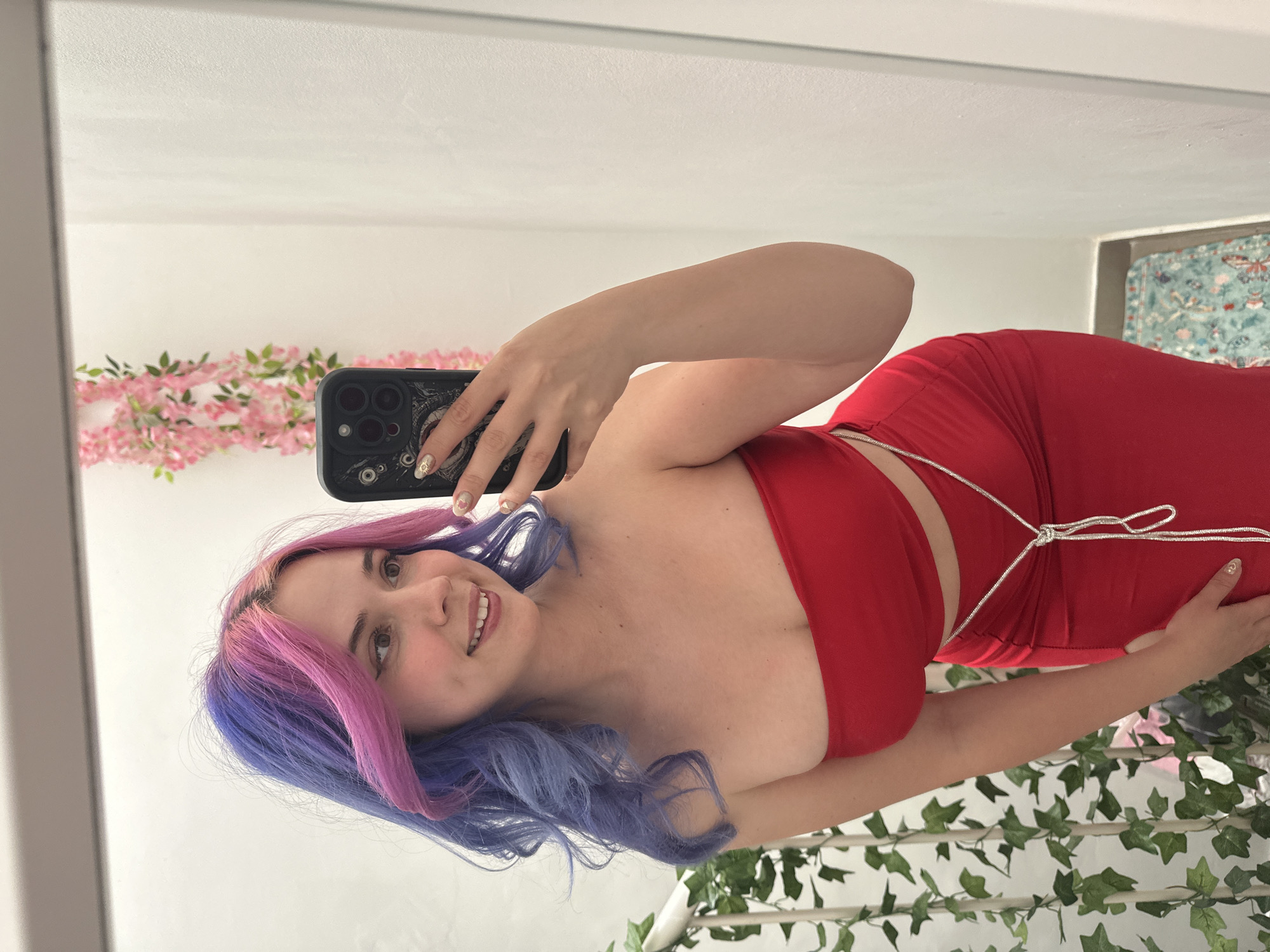 A young woman with vibrant purple and pink dyed hair is captured in a mirror selfie, smiling warmly at the camera. She is wearing a form-fitting, strapless red two-piece outfit, consisting of a crop top and a high-waisted bottom. A thin, shiny white cord is tied around her midriff, connecting or accentuating the outfit. She holds a dark-colored smartphone with multiple camera lenses in her right hand, which also displays subtle nail art.
The setting appears to be an indoor space, likely a bedroom or dressing area, as suggested by the reflection in the mirror. In the background, to the left, a string of pink artificial flowers or foliage is visible against a white wall. To the right and lower portion of the reflection, a green leafy garland or artificial ivy is draped. A section of wall art or a tapestry with a light blue background and butterfly patterns is partially seen in the upper right corner of the reflected space. The overall impression is one of a casual, posed portrait. No visible text is legible in the image.