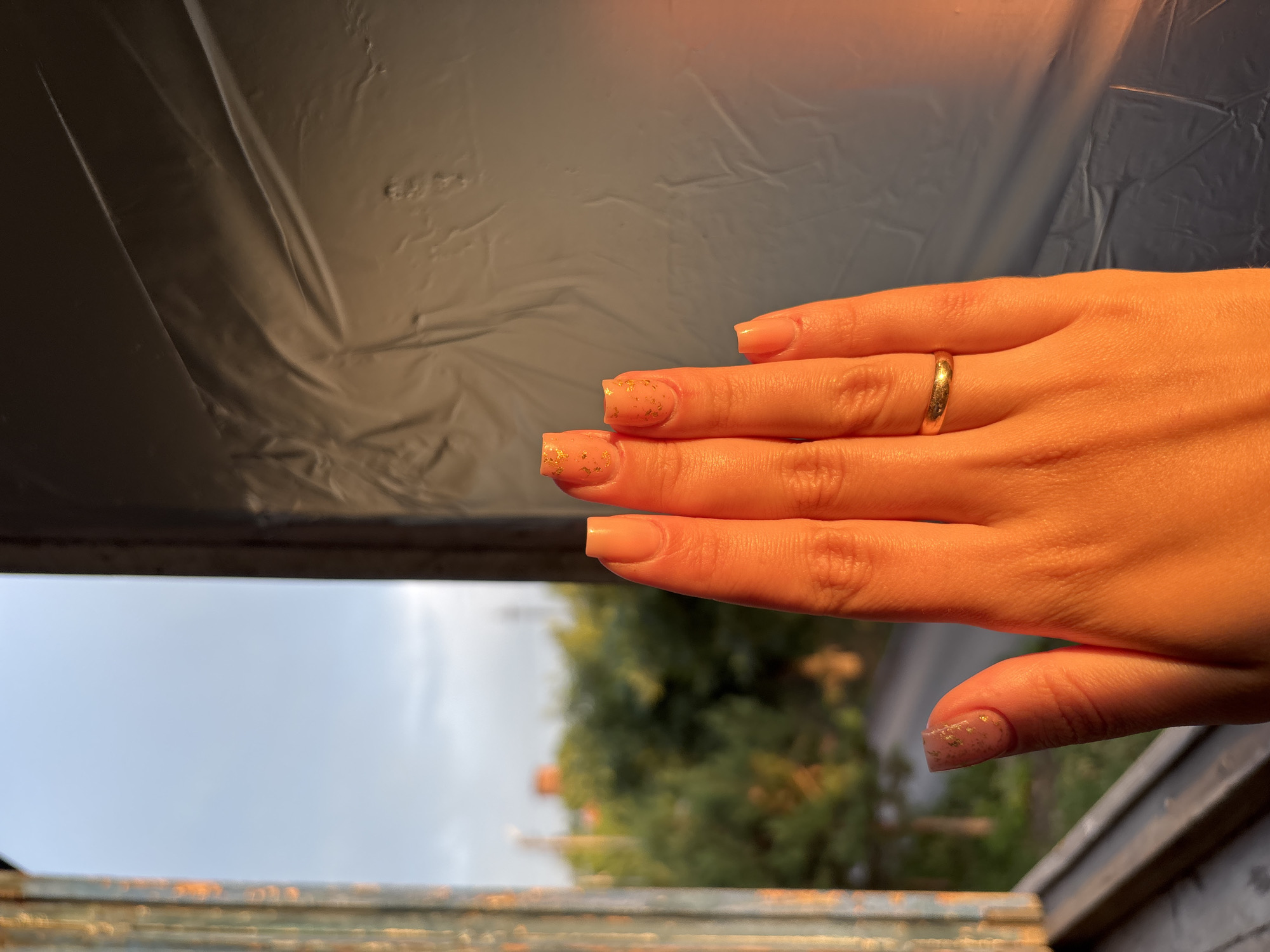 A close-up, eye-level shot focuses on a person's left hand, which is illuminated by warm, golden sunlight, suggesting it is either sunrise or sunset. The hand, with manicured nails painted a pale orange and adorned with gold flecks, is held up against a background that includes a window frame, blurry trees and buildings, and a textured, gray surface on the right. A simple gold band is visible on the ring finger. The overall impression is of a relaxed, contemplative moment, possibly taken at a residence in Inskoy, Russia, given the presence of greenery and building structures often found in urban or suburban environments.