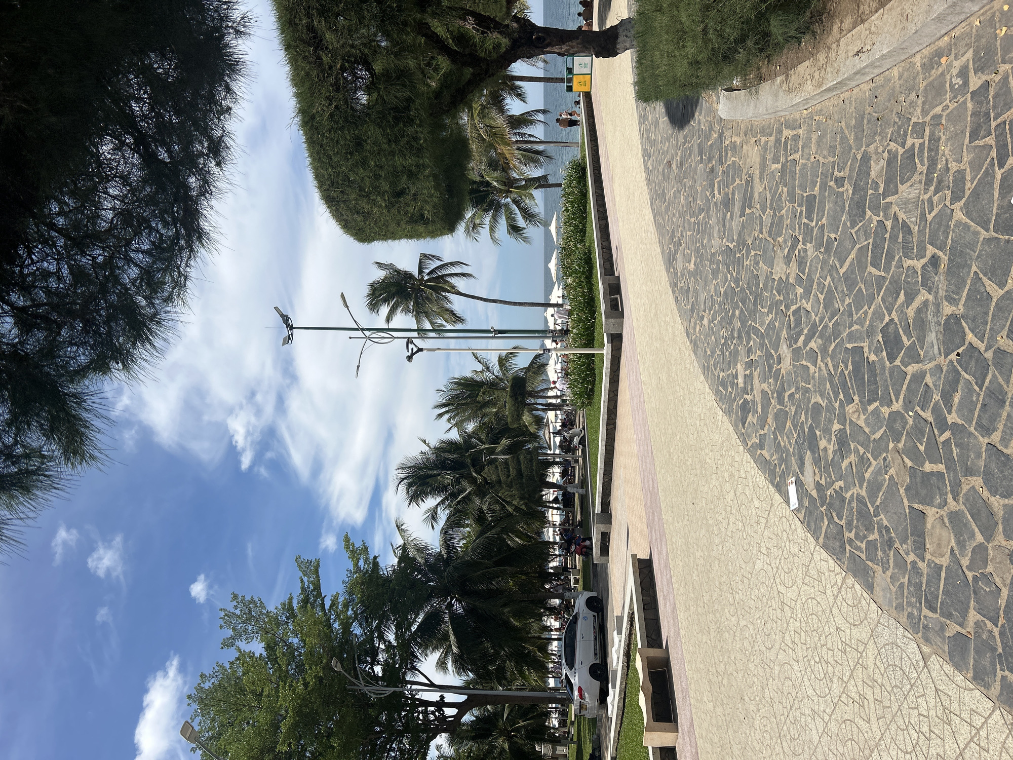 The image depicts a paved walkway on a sunny day, likely in Nha Trang, Vietnam, overlooking a beach. The walkway is paved with a mosaic of dark gray stones in the foreground, curving to the right. To the left, a lighter patterned pavement features circular motifs. Several benches made of concrete and stone are visible along the edge of the walkway.
Beyond the walkway, a green grassy area with palm trees and lush vegetation leads to the beach. In the middle ground, a white sedan is parked. Further back, a line of white umbrellas indicates a beach seating area. People are seen strolling on the beach and near the water. The ocean is visible in the background under a bright blue sky with scattered white clouds. Tall streetlights are positioned along the walkway. The overall scene is serene and suggests a popular tourist destination.