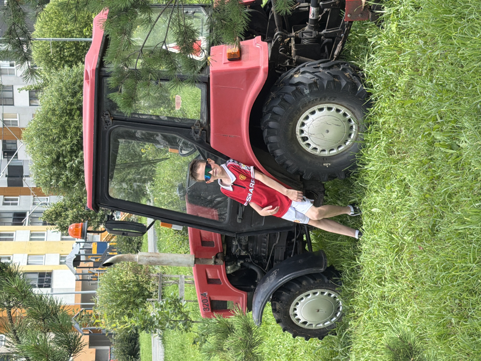 A young boy wearing a red soccer jersey and white shorts with sunglasses on his face is leaning against a red tractor parked on a grassy lawn. The tractor is a Belarus model, with the number "320.4" visible on its side. In the background, there are multi-story apartment buildings and playground structures, suggesting an urban residential area. The presence of pine branches in the foreground indicates a possible park or a residential area with landscaping. The scene appears to be outdoors during the daytime, with bright natural light. The location is Yekaterinburg, Russia.