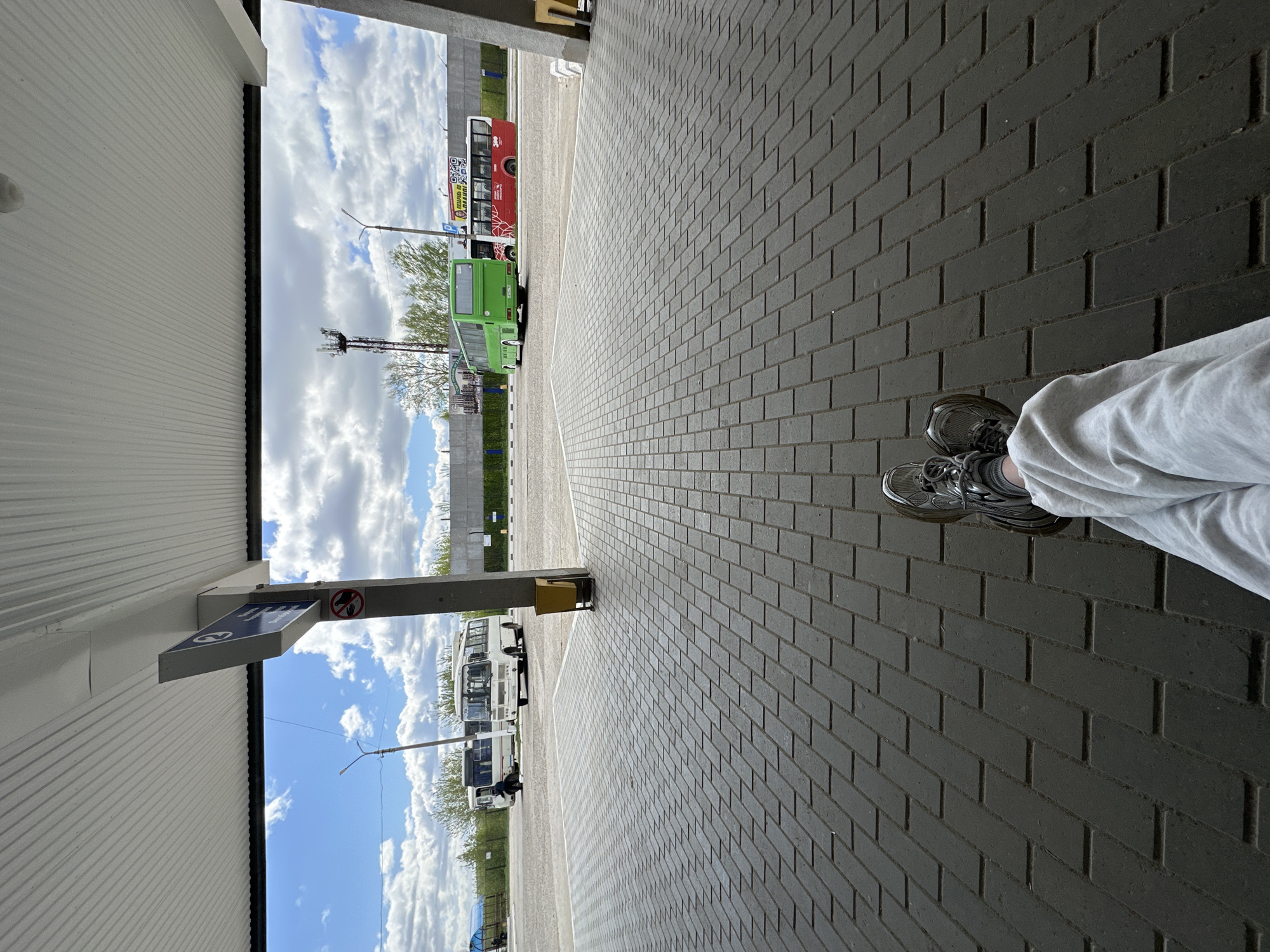 The image depicts a ground-level perspective of a paved area, potentially a bus station or waiting area, in what appears to be Chaikovsky, Russia. The ground is covered with gray brick pavers arranged in a repeating pattern. To the left, there's a partially visible corrugated metal structure, likely a shelter or a building facade. A blue and white sign with symbols is attached to the structure, including a no-smoking symbol.
In the background, several buses are parked. A green bus and a red bus with a prominent sign are visible in the mid-ground, further back are multiple white buses. A cell tower can be seen on the left. The sky is visible in the background, with a mix of blue sky and scattered clouds. The overall scene seems to be taken on a partly cloudy day.
In the right corner of the image, a person's legs are visible, clothed in light gray pants, with silver-colored sneakers on their feet. They appear to be sitting or reclining while taking the photograph.
