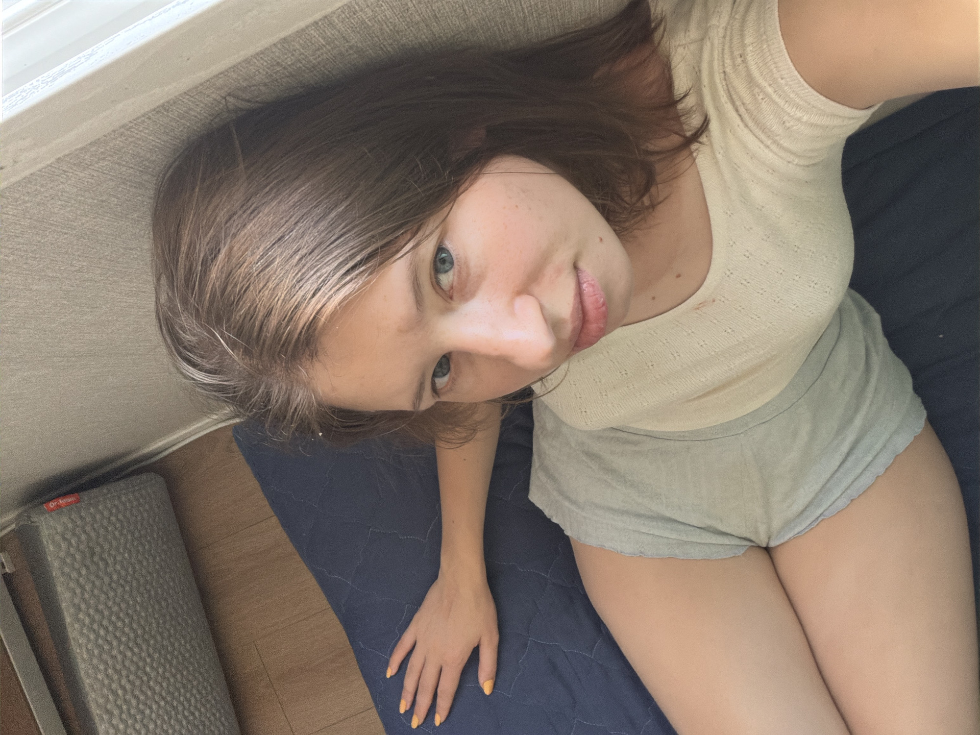 A young woman with long brown hair and blue eyes is lying on a blue mattress in what appears to be a bedroom. She is wearing a cream-colored short-sleeved top and light blue shorts. Her left arm is extended, resting on the mattress, and her fingernails are painted yellow. In the background, a grey yoga mat is visible on a wooden floor, and a textured wall is to the right. The overall lighting suggests it is daytime, with light coming from a window on the right side of the frame. The location is identified as Dongducheon-si, South Korea.