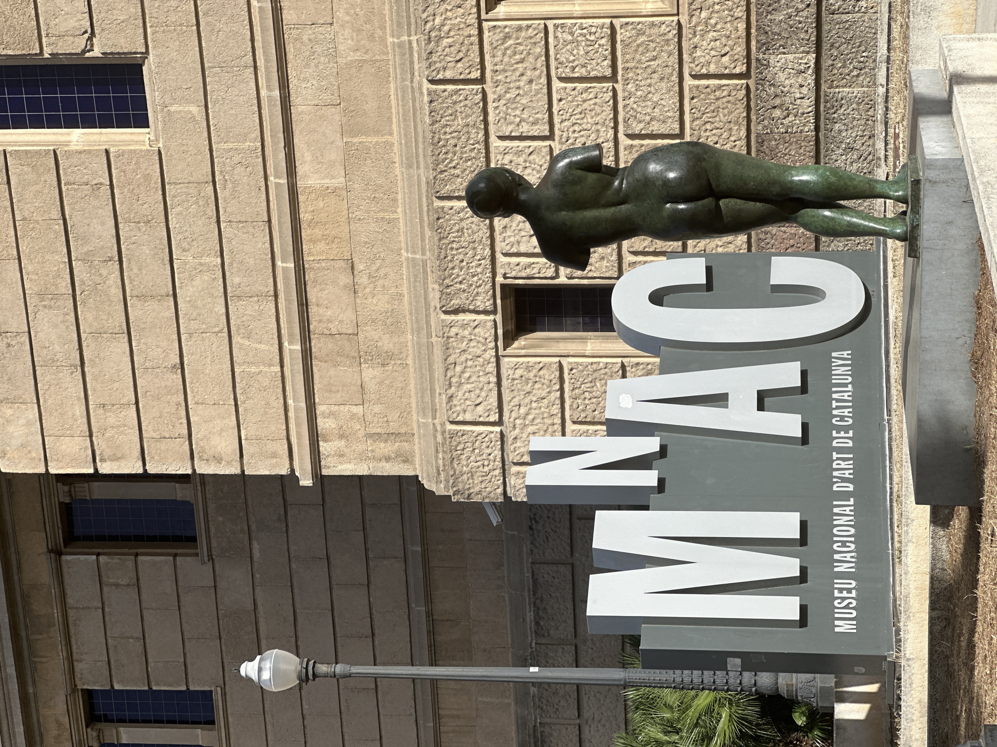 The image shows a bronze statue of a nude female figure, viewed from the back, standing in front of large, three-dimensional letters that spell out "MNAC". Below these letters, the text "MUSEU NACIONAL D'ART DE CATALUNYA" is visible. The statue is positioned to the right of the letters, with its back towards the viewer and its head slightly turned to the left. The letters are light gray and appear to be made of metal or a similar material, mounted on a dark gray base.
The background is a building constructed of light brown stone blocks, with dark windows and architectural embellishments. To the left of the letters, a lamppost with a white, globe-shaped light fixture is visible. Palm fronds from a plant can be seen in the lower left corner. The ground in front of the display is covered in dry grass and leaves. The scene suggests an outdoor setting in front of a prominent cultural institution. This is the entrance to the Museu Nacional d'Art de Catalunya (National Art Museum of Catalonia) in Barcelona, Spain, located on Montjuïc hill.