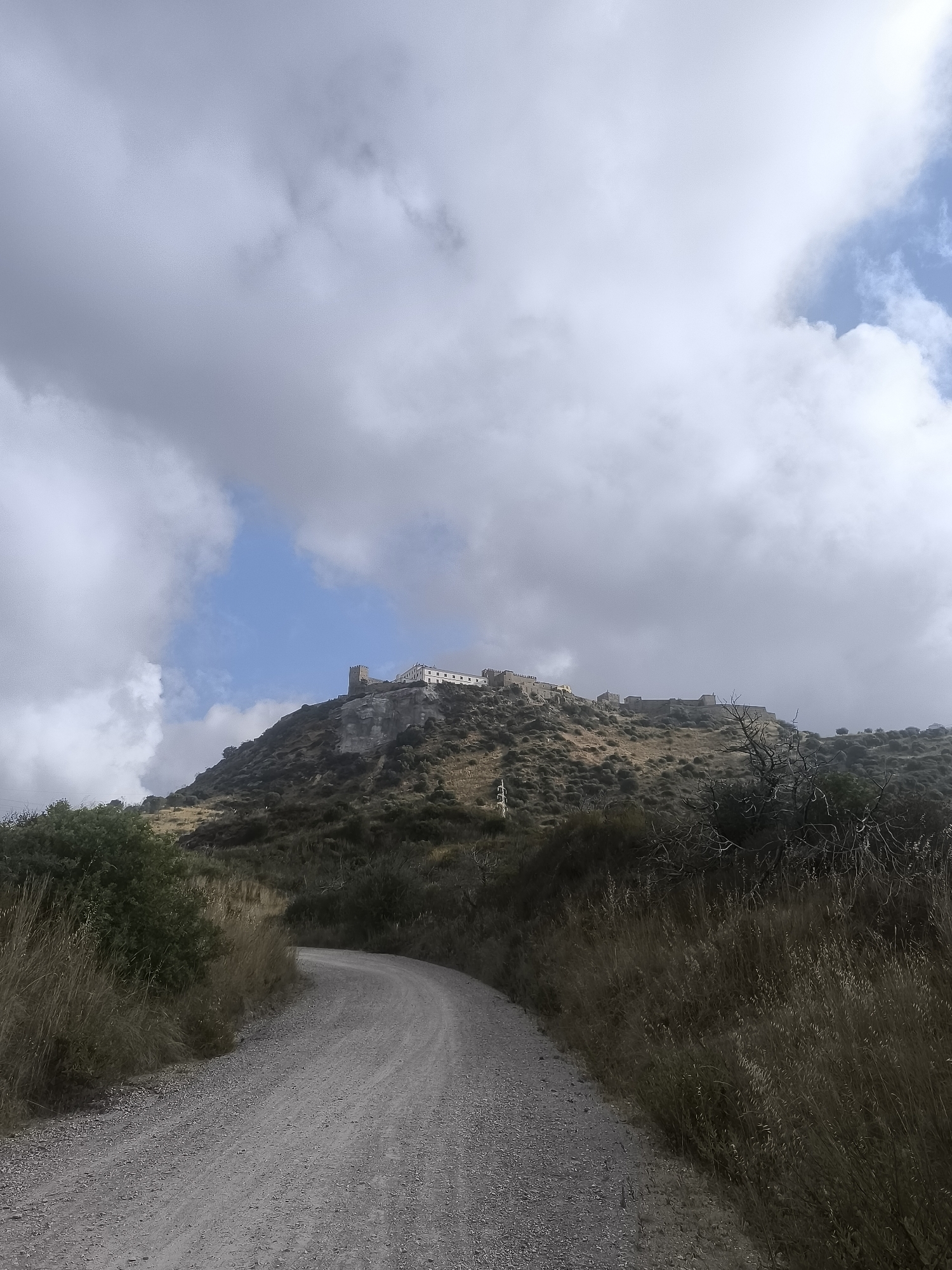 A dirt road curves towards the viewer from the bottom left of the frame, disappearing around a bend. The road is flanked by dry grass and scrubby bushes. In the distance, atop a hill, is a white castle complex with a few stone towers. The sky above is filled with dramatic, grey clouds, with patches of blue sky visible.