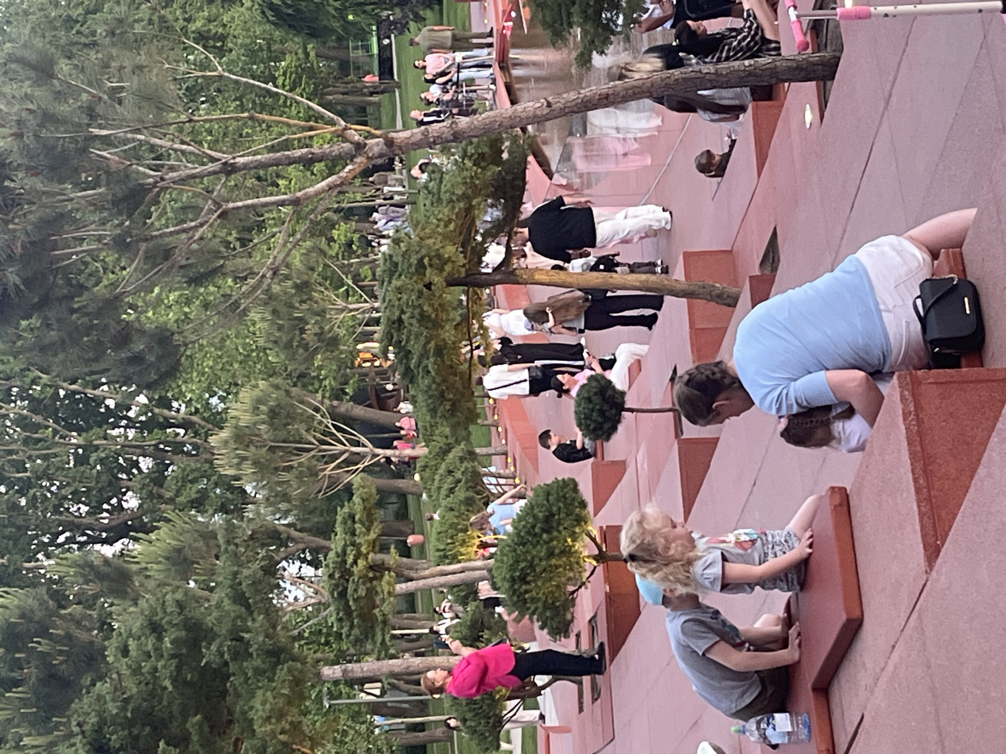 An outdoor scene captures a public park or plaza during what appears to be late afternoon or early evening, characterized by soft, natural light. The image is oriented sideways, with architectural elements forming the foreground on the right and a lush park extending towards the left background.
In the immediate foreground, a woman in a light blue shirt and white shorts is leaning forward, looking down, with a black handbag resting beside her. Adjacent to her, a young child with blonde hair, wearing a grey t-shirt and patterned shorts, sits on a distinctive red-brown terraced surface. Below, a boy in a grey t-shirt also sits on a lower terraced level, with a plastic water bottle nearby.
The midground features more of these red-brown, stepped architectural structures, which appear to be part of an outdoor seating or viewing area. Small, round topiary trees are strategically placed on these platforms. A woman in a bright pink jacket and black pants stands here, holding a smartphone, seemingly capturing a photo or video. Numerous other people, including adults and children, are scattered across the various levels and pathways, some standing, others walking or observing. To the left of the central structures, a calm body of water or pond reflects the sky and surrounding trees.
Further into the background, the scene transitions into a dense green park or garden area, filled with various trees, some with prominent branches, others forming a thick canopy of foliage. More distant people are visible, enjoying the outdoor environment. The overall atmosphere suggests a leisurely public gathering or stroll in a well-maintained recreational space, consistent with a park or promenade in a city like Krasnodar, Russia. No visible text is clearly legible.