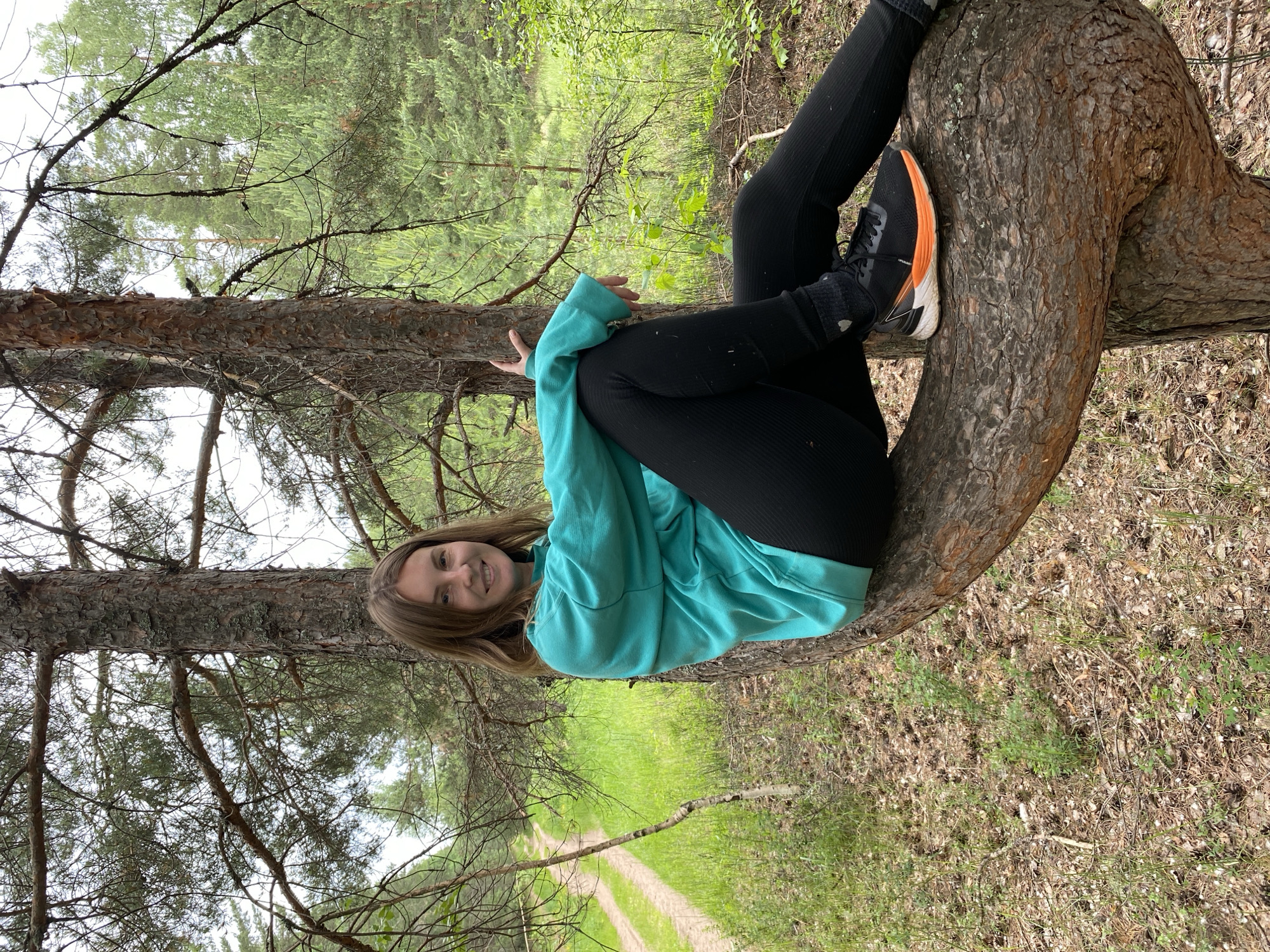 A young woman is pictured sitting on a thick, curved tree branch in a forest. She is wearing a bright teal hooded sweatshirt and black leggings, with black and orange athletic shoes. Her legs are bent, and one foot rests on the branch. She is smiling at the camera. The tree branch is rough and textured, with bark visible. The forest setting includes pine trees with needles and a dirt path visible in the background. The lighting suggests it is daytime, possibly overcast. The overall mood is casual and happy.
