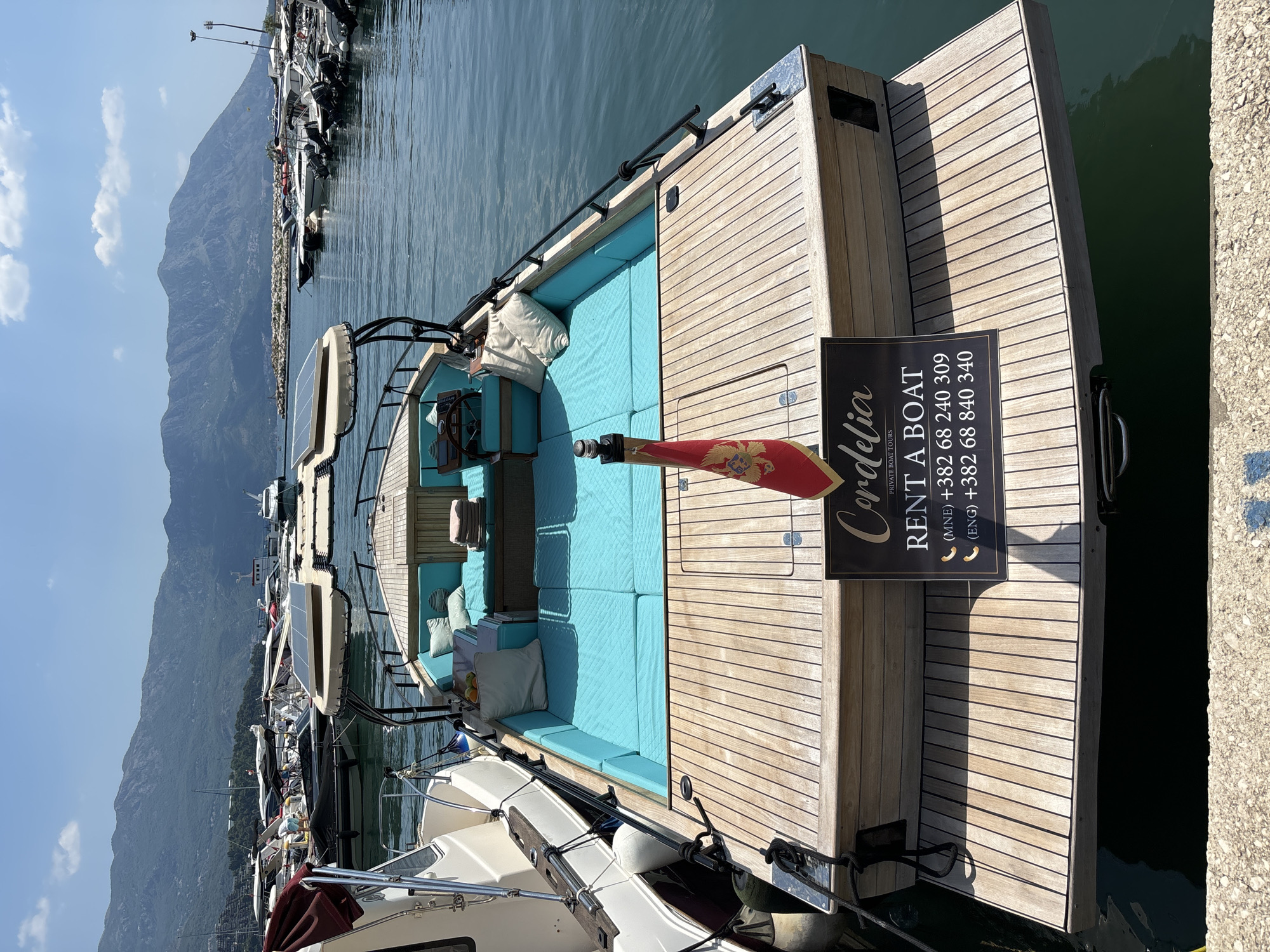 This media file captures a close-up, eye-level view of the stern of a wooden boat docked in a harbor. The boat features light turquoise upholstered seating and several white cushions. A small flag with a red background and a golden crest is prominently displayed.
To the right of the boat, a black sign with gold lettering reads "Cordelia PRIVATE BOAT TOURS RENT A BOAT" followed by phone numbers listed as "(MNE) +382 68 240 309" and "(ENG) +382 68 840 340". The wooden decking of the boat is well-maintained and shows the distinct grain of the wood.
In the background, the water of the harbor is visible, reflecting the clear blue sky. The scene is set against a backdrop of a large, mountainous landscape, suggesting a coastal or fjord-like location. Other boats are docked nearby, indicating a bustling marina. The lighting suggests it is a sunny day, casting shadows on the boat's deck. The overall impression is one of a luxurious and well-appointed vessel available for hire in a picturesque setting, likely Budva, Montenegro, given the phone number prefixes.