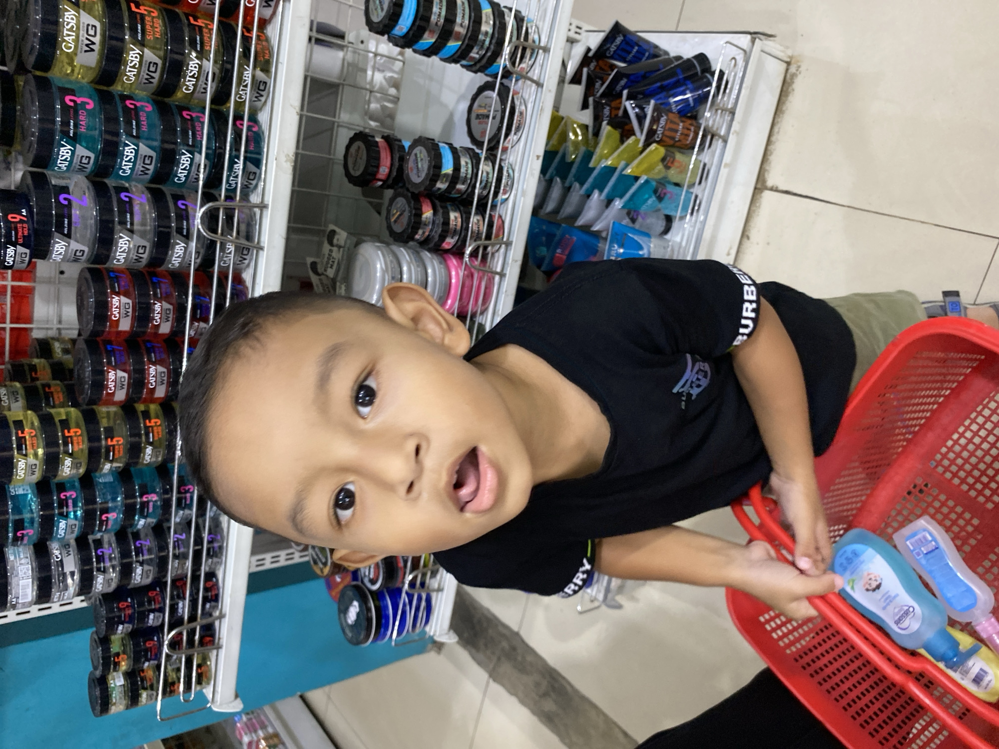 A young boy is standing in an aisle of a store, holding a red basket. He is looking directly at the camera with his mouth open. He is wearing a black t-shirt and khaki shorts. The shelves behind him are stocked with various hair styling products, many of which are branded "Gatsby". The store appears to be a typical retail environment with tiled flooring. The provided crops indicate the location is in Lampisang, Indonesia.