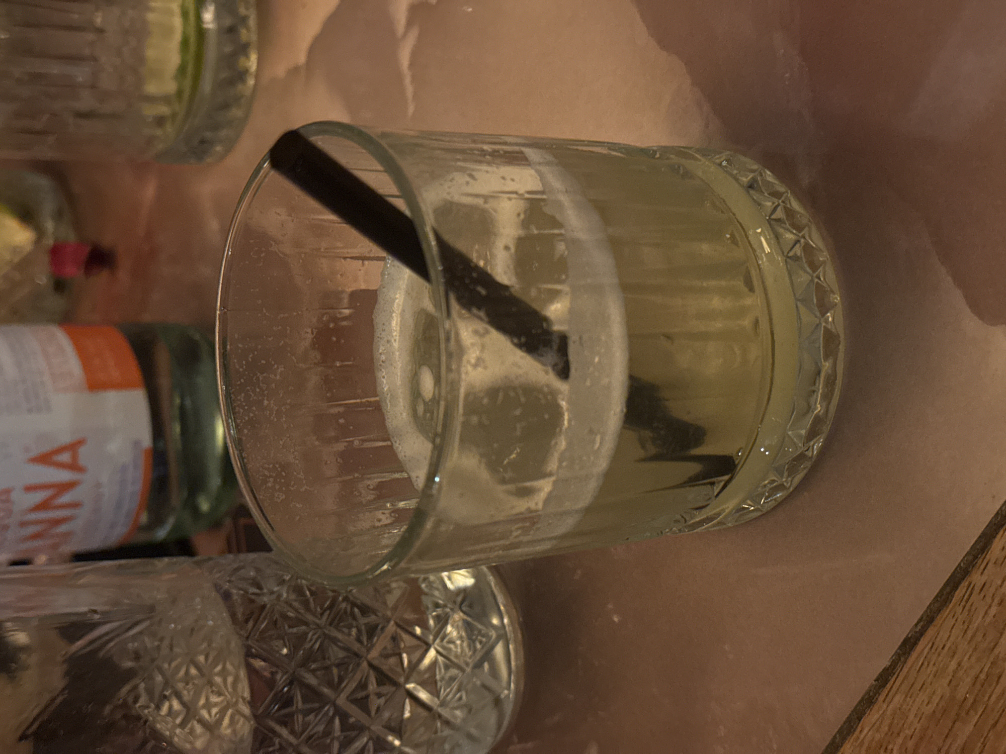 The image shows a partially consumed cocktail in a faceted glass with a black straw, sitting on a textured surface that appears to be a table. To the left, blurred in the background, are other glasses and a bottle labeled "ANNA." The context clues suggest a restaurant or bar setting. There is no information available to determine whether the image was taken in the city of Sofia, Bulgaria.