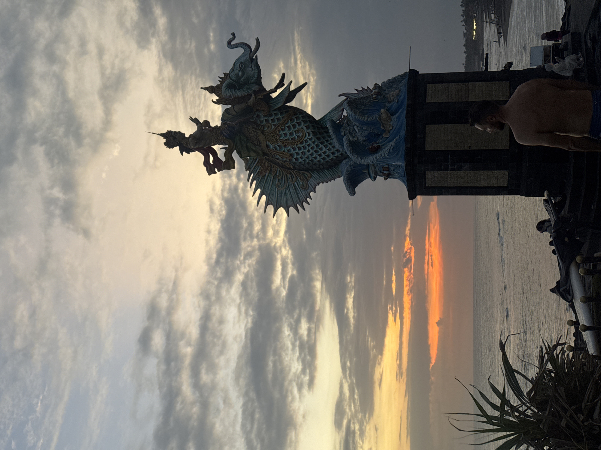 At sunset in Mengwi, Indonesia, a large, ornate statue of a mythical creature resembling a fish with an elephant riding on its back stands prominently. The statue is supported by a dark, multi-tiered base. A man, shirtless and wearing blue shorts, stands with his back to the camera, looking out at the ocean. The sky is filled with dramatic clouds, transitioning from soft yellow and orange hues near the horizon to darker gray tones above. The ocean is visible in the background, with gentle waves rolling towards a dark sand beach. Several other people are visible in the distance on the beach. Lush green vegetation is present in the foreground to the left of the frame.