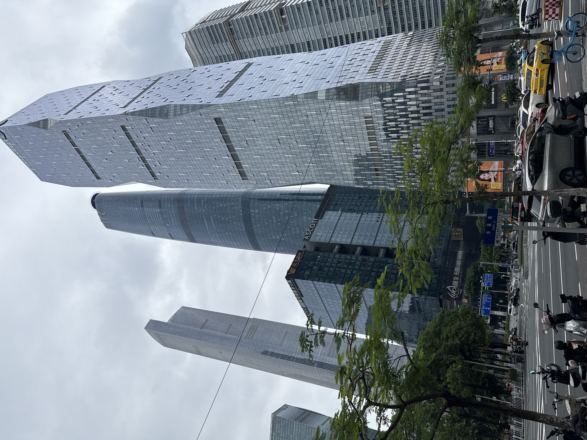 The image depicts a street view in Tianhe District, China, characterized by a cluster of modern skyscrapers under an overcast sky. In the foreground, a street with multiple lanes of traffic is visible, with numerous parked and moving vehicles including cars, motorcycles, and a blue bicycle. Several trees line the sidewalk on the left and right, partially obscuring the lower sections of the buildings. Signs for businesses such as "Ascott," "McDonald's," and "iFC shopping mall" are discernible on the lower facades of the buildings. The architecture of the skyscrapers is varied, with some featuring sleek, glass-covered exteriors and geometric designs, while others have more traditional rectangular forms. The overall scene suggests an active urban environment.