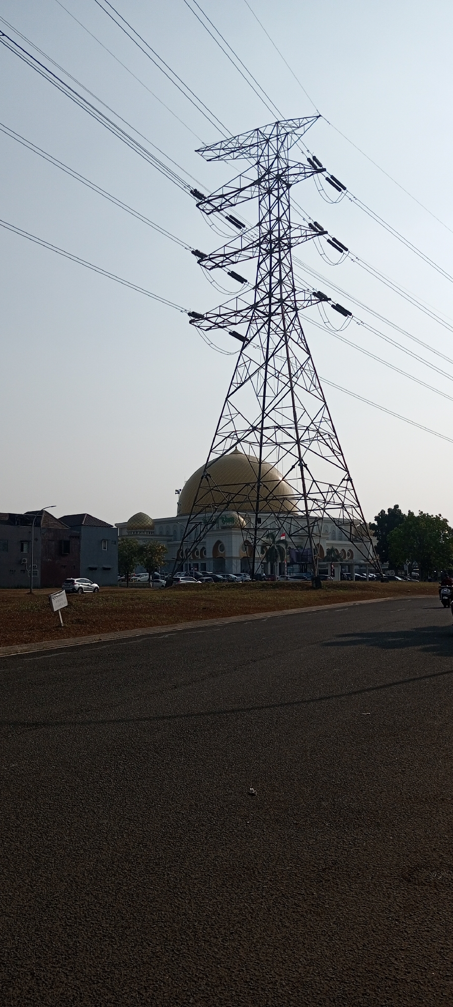 In Lambangjaya, Indonesia, an immense steel electricity pylon dominates the foreground, its complex lattice structure silhouetted against a pale, cloudless sky. Multiple high-voltage power lines stretch across the frame, emanating from the pylon and disappearing towards the horizon. Behind the pylon, the golden dome of a mosque gleams, its architectural features partially obscured by the pylon's framework. Surrounding the mosque are several buildings and a parking lot filled with various vehicles. A paved road occupies the bottom portion of the image, with a grassy area and a small sign to the left. The overall scene depicts a blend of industrial infrastructure and religious architecture under a clear sky.