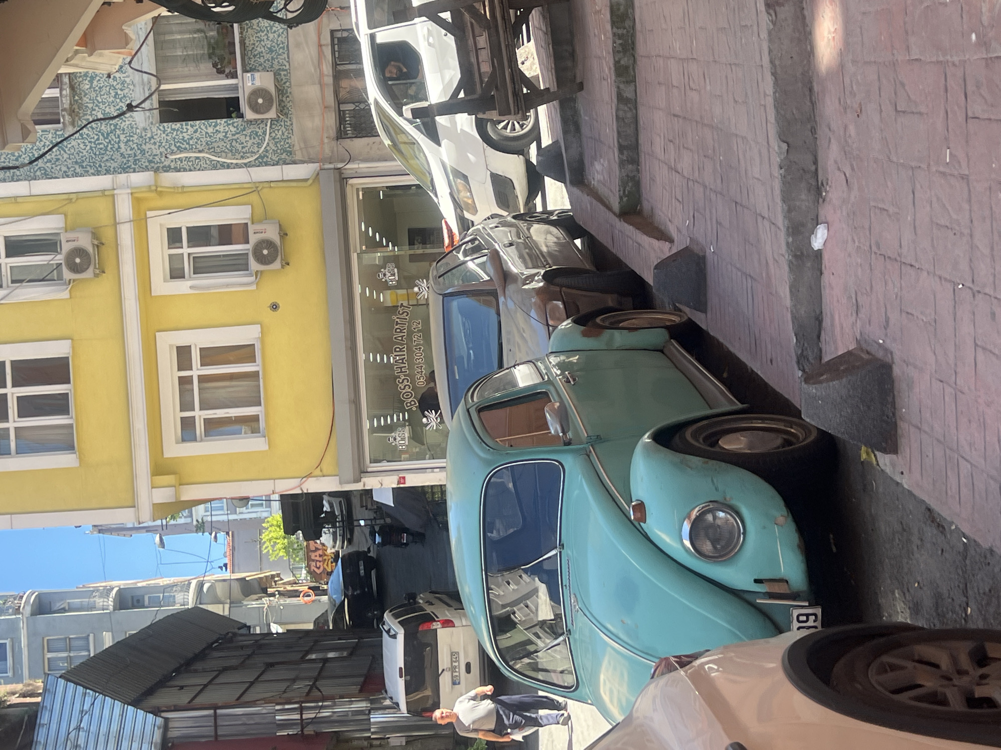 The image captures a street scene in Beyoğlu, Turkey. Parked cars are visible along the street, including a light blue vintage Volkswagen Beetle. A person in a gray shirt and dark pants walks near the cars. Buildings line the street, one of which is a yellow building with white-framed windows and air conditioning units. A business called "BOSS HAIR ARTIST" is identifiable. The street is paved with red bricks. Overhead, there are utility lines and architectural details of the buildings.