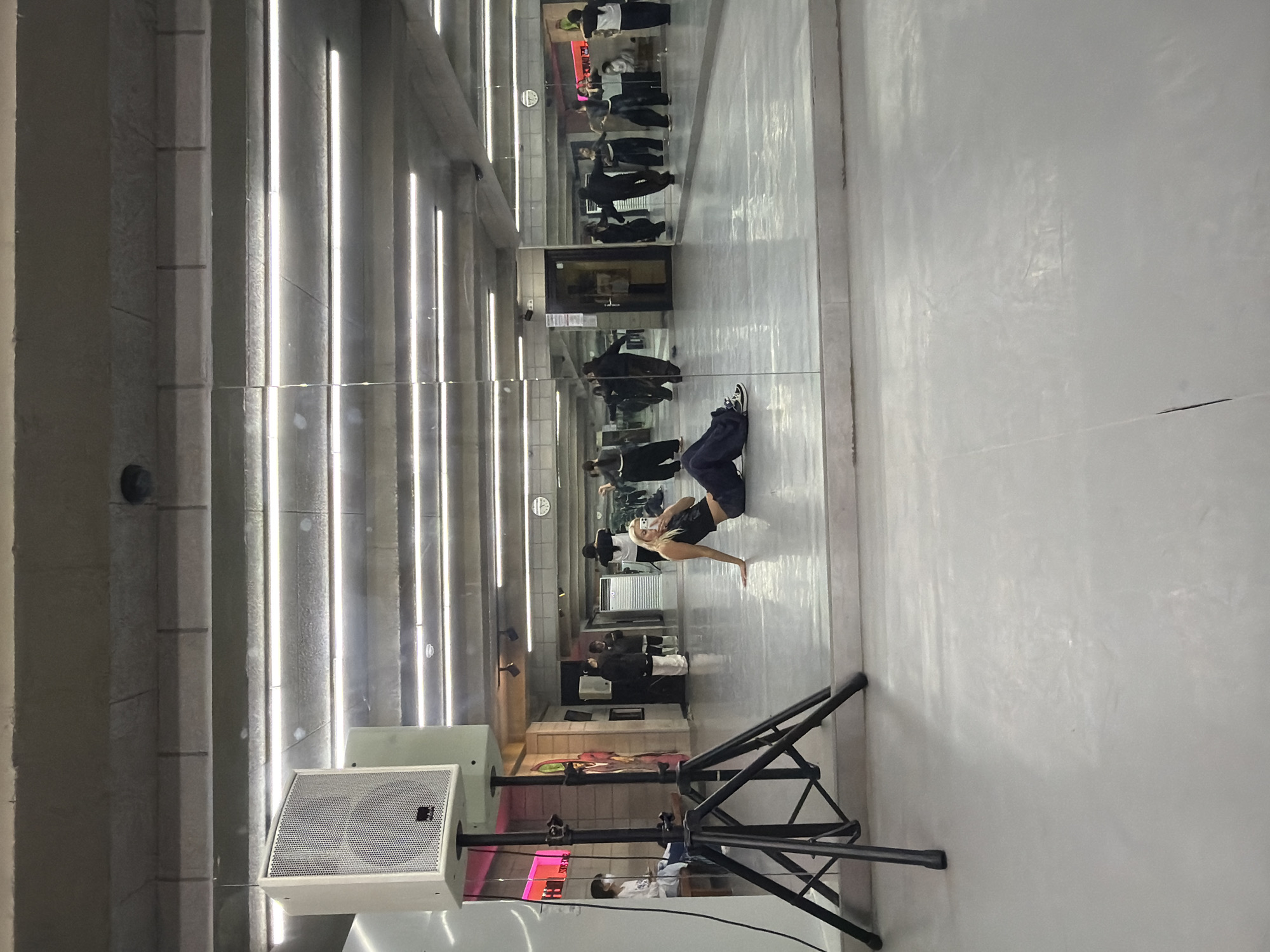 A long shot photograph reveals a vertical orientation that captures an indoor dance studio environment. A woman wearing dark clothing is centered in the frame, engaged in a breakdancing pose. She's balanced with one hand on the floor and her body arched. The scene is reflected in a mirror that stretches the length of the room. In the mirror, additional people can be seen, although their actions are not as clear. The background features a long corridor lined with mirrors. The corridor is brightly lit with rows of overhead lights. In the foreground, on the left side of the frame, is a large white speaker mounted on a black metal stand. The speaker dominates this section of the photograph. This is in Seoul, South Korea.