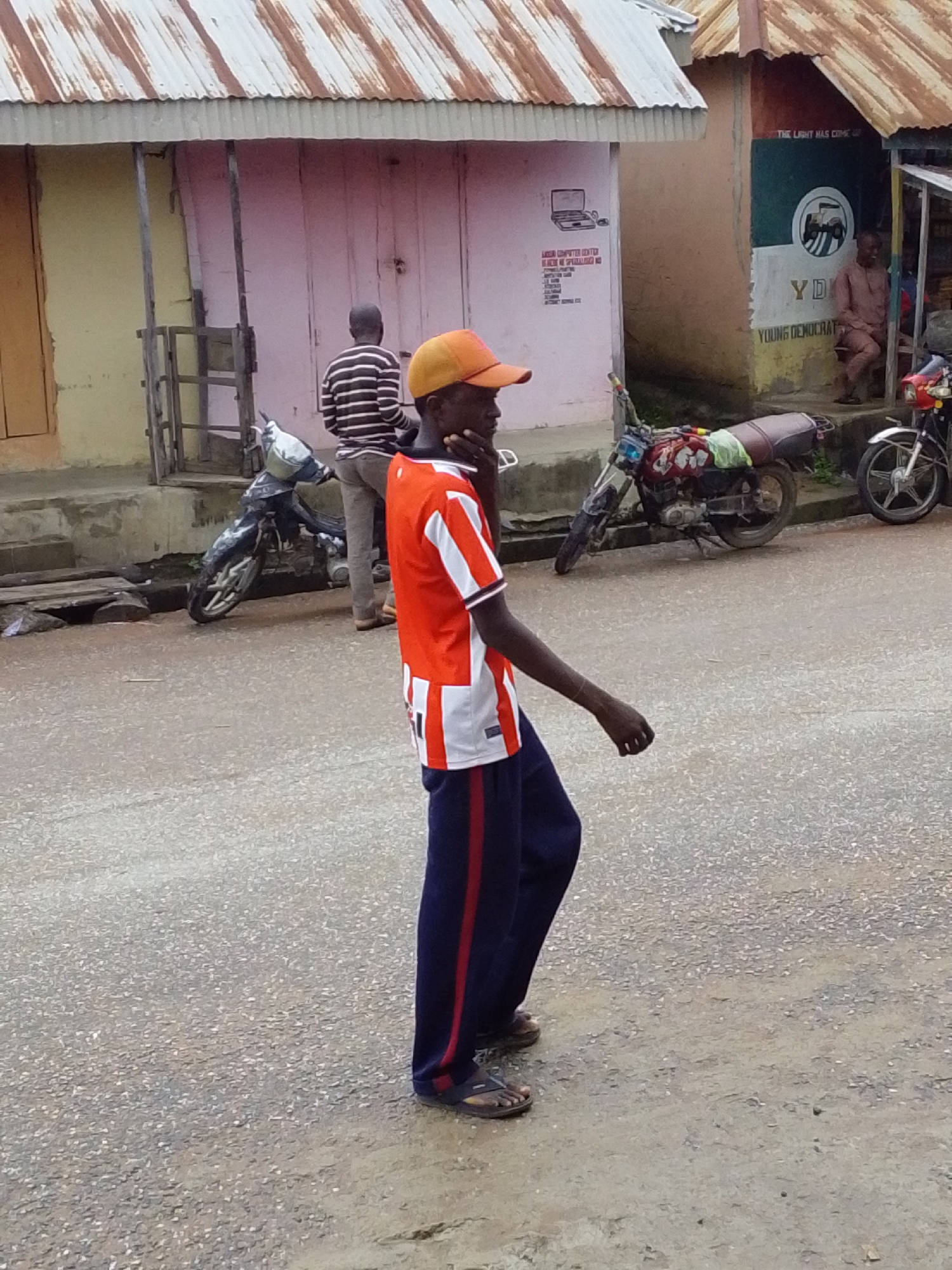 A young man wearing an orange and white striped t-shirt and dark blue pants with red stripes is standing on a wet asphalt road in front of a light yellow building with a corrugated tin roof. He is looking to his left, with his left hand raised to his face and his right arm extended to his side. His feet are clad in brown and black sandals. Behind him, a black motorcycle with a red and white patterned tank is parked on the street. To the left of the motorcycle, another motorcycle is partially visible. Further back, a man in a black and white striped shirt stands near a doorway. To the right of the yellow building, a pink-colored structure with a mural depicting a tractor and a green circular emblem is visible. A man in a brown outfit is seated near this structure. The scene appears to be a street in Serti, Nigeria, characterized by its local architecture and everyday street life.