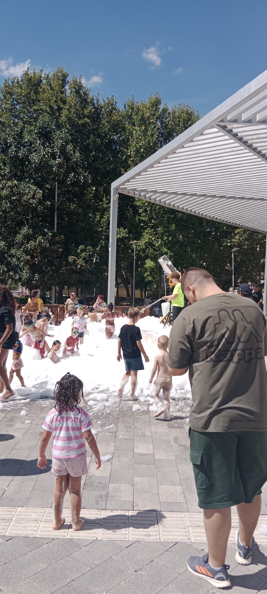 The image depicts an outdoor foam party in Alcalá de Henares, Spain, likely during the summer. The scene is set under a white modern canopy structure, with lush green trees and a clear blue sky visible in the background. A foam machine is actively spraying foam into a large pool filled with children. Several children are playing in the foam, some partially submerged, while others are splashing and interacting. Adults are present on the periphery, supervising the activity. One adult in the foreground is wearing a green t-shirt with a Kappa logo. The ground is paved with gray and beige square tiles, with some foam scattered on it. The overall atmosphere appears to be one of fun and recreation for children.