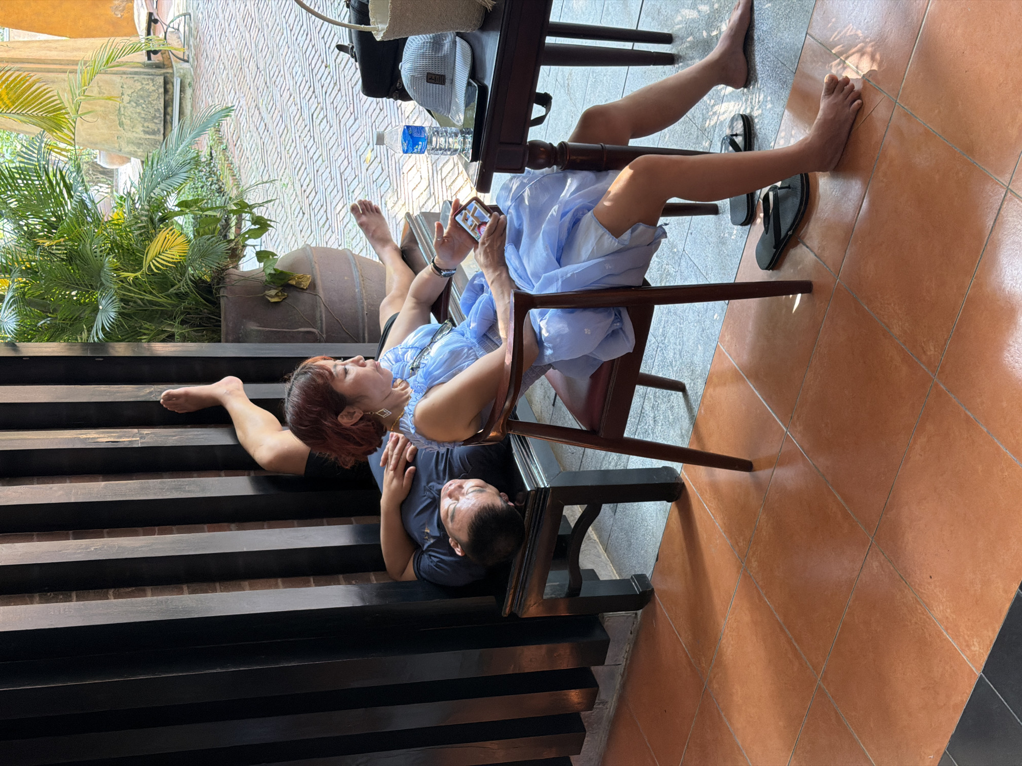 The image shows a middle-aged couple in an outdoor setting. A woman with reddish-brown hair and a blue dress sits on a dark wooden chair, looking at a smartphone in her hands. She wears jewelry. Her bare feet are resting on a dark wooden structure resembling steps. A man in a dark t-shirt is lying on the steps with his head resting near the chair, his hands near her body. Parts of both people's legs and feet are visible. A table is nearby with a water bottle and other items. The ground is covered with tiles. In the background, there are plants in a pot and a brick wall. Based on architectural elements, plants, and styling, the location could be a cafe or restaurant setting in Đà Nẵng, Vietnam.