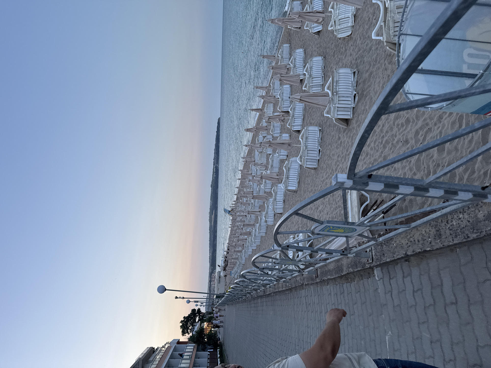 The image depicts a view of a beach in Obzor, Bulgaria, during the late evening or early morning. The foreground features a paved walkway with a metal railing separating it from the sandy beach. The beach is lined with numerous rows of empty lounge chairs and closed umbrellas, suggesting the end of the day or the beginning of the season. A building, possibly a hotel, is visible on the left side of the walkway. The sea stretches out in the background under a clear, pale sky. A few people are walking along the walkway in the distance.