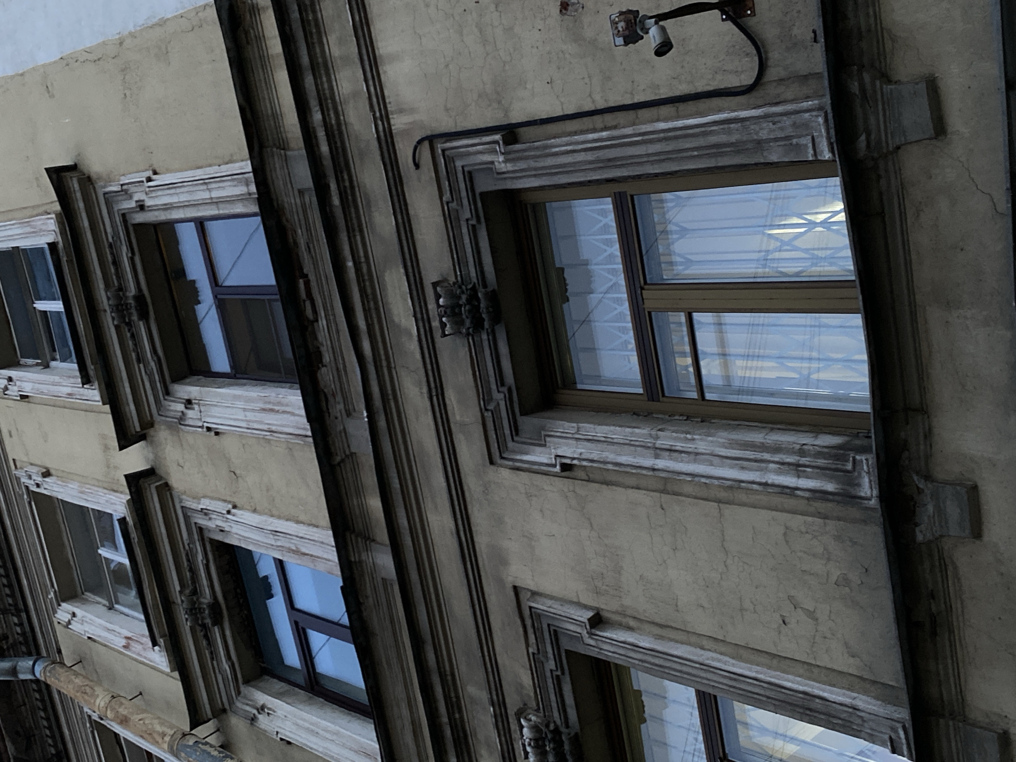 The image depicts a building exterior, likely in Saint Petersburg, Russia, characterized by its weathered facade and architectural details. Several windows are visible, each framed with ornate molding. A surveillance camera is mounted on the wall above the uppermost window. The scene is shot from a low angle, looking upwards, emphasizing the height of the building. A drainpipe runs vertically along the wall. The overall color palette is muted, with grays and browns dominating.
