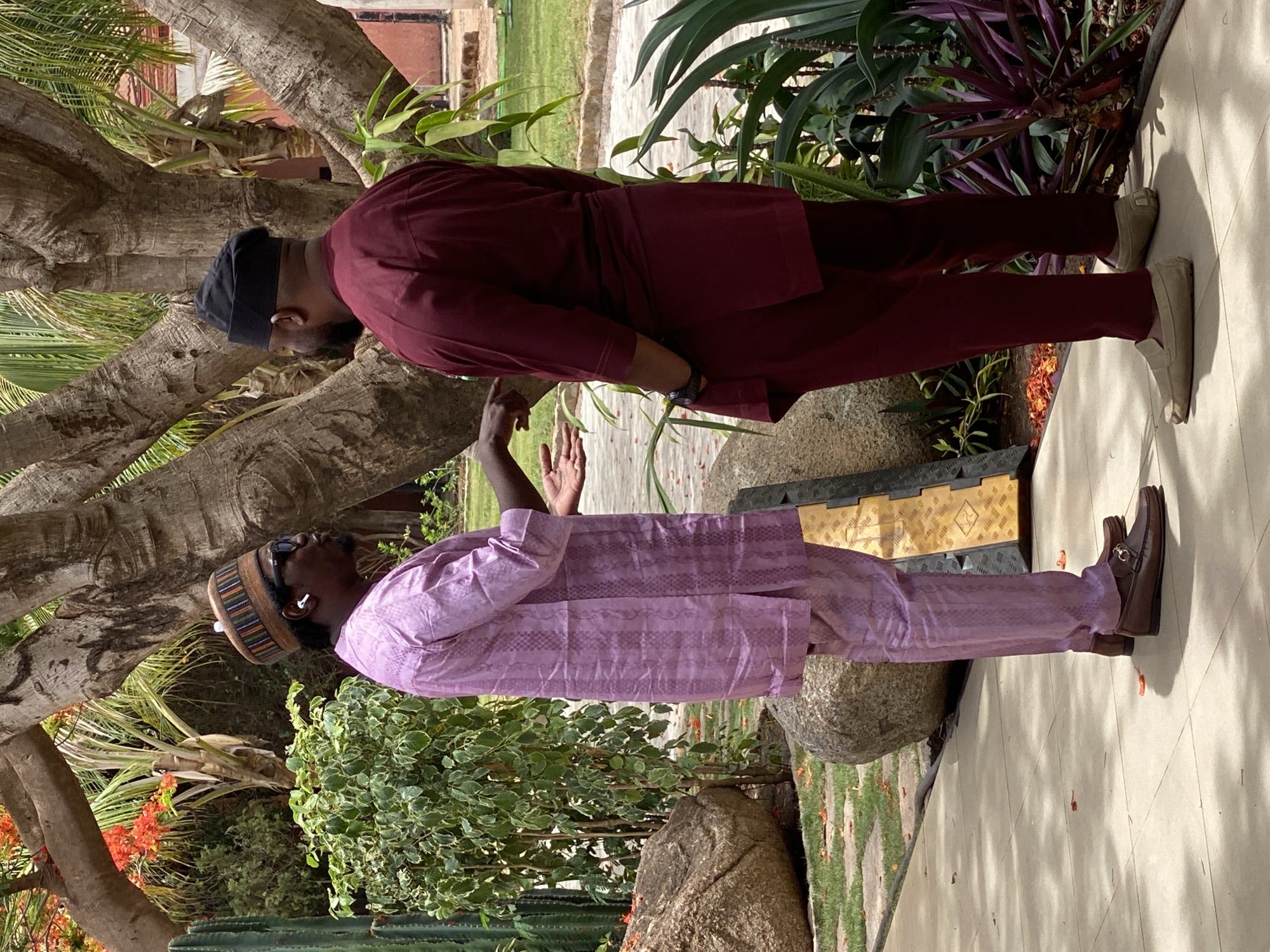 The image shows two men engaged in conversation outdoors, likely in a garden or courtyard setting in Kwarau, Nigeria, under bright daytime conditions.
The man on the left wears a lavender-colored traditional outfit with a subtle pattern, a multi-colored striped cap, and dark sunglasses. White wireless earbuds are visible in his ears. He stands facing the right, with his right arm raised and hand open, seemingly mid-gesture during a discussion. He is wearing brown loafers with a metallic buckle.
The man on the right, dressed in a maroon traditional outfit and a black skullcap, stands facing the left. He appears to be looking down towards the other man's gesturing hand or the ground. He wears light-colored, possibly beige, slip-on shoes.
The setting is lush with abundant greenery, featuring large trees with rough bark, tropical fronds, and various green and purple leafy plants. Several large rocks are scattered on the ground, and a yellow and black textured mat or cable protector is visible near the man in lavender. Both men are standing on a light-colored paved surface, with noticeable shadows indicating strong sunlight. No discernible text is visible in the image.