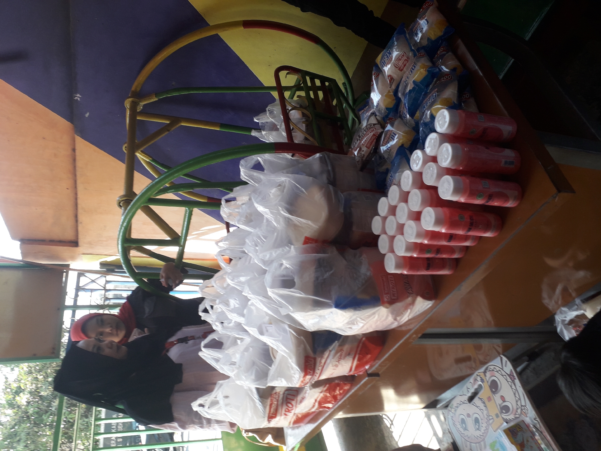 In Cankuang, Indonesia, two women, one wearing a black hijab and the other a red hijab, stand behind a table laden with grocery bags and packaged goods. The woman in the black hijab is looking to her left, while the woman in the red hijab is looking forward, her left hand resting on a colorful playground swing behind them. The swing, a circular structure painted green and yellow with red accents, is positioned behind the table. The table itself is an orange wooden surface, on which are arranged numerous white plastic grocery bags, some appearing to contain food items like bread. Several small, pink bottled beverages are also neatly lined up on the table. In the background, a wall painted with diagonal bands of purple and yellow provides a backdrop. Sunlight streams in from the left, casting shadows and highlighting the scene. A small, child's activity table with cartoon drawings is visible on the left side of the image.