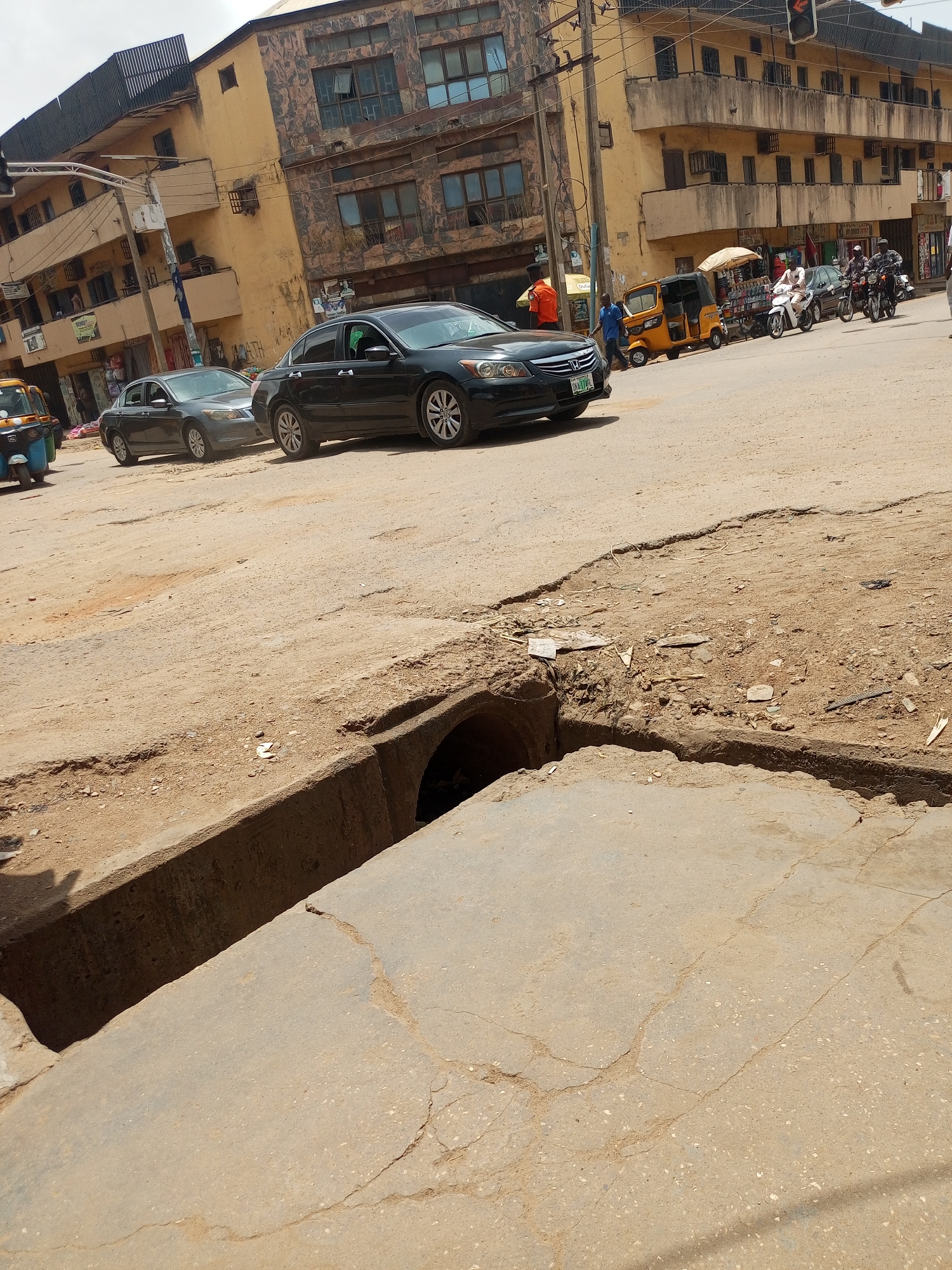 The image depicts a street scene in Maiburiji, Nigeria. In the foreground, a large open drainage ditch runs across the road, posing a hazard. The road surface is cracked and uneven. Further back, two dark-colored cars are visible, as well as several motorcycles and auto rickshaws. Buildings line the street, and pedestrians are visible in the background. A traffic light hangs overhead. The scene appears to be a busy intersection.