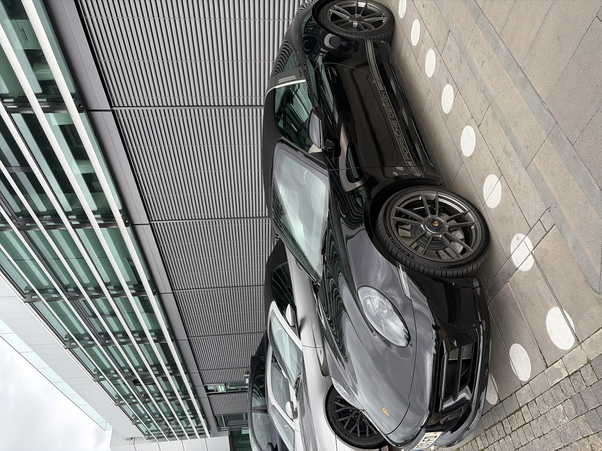A black Porsche Design Edition parked on a sidewalk in Tbilisi, Georgia, is photographed from a low angle. The car is situated next to a modern building featuring alternating bands of glass and metal louvers. The sidewalk is paved with gray cobblestones and marked with white circular indicators. A second, lighter-colored car is visible behind the Porsche. The photograph is taken during daylight under overcast conditions.