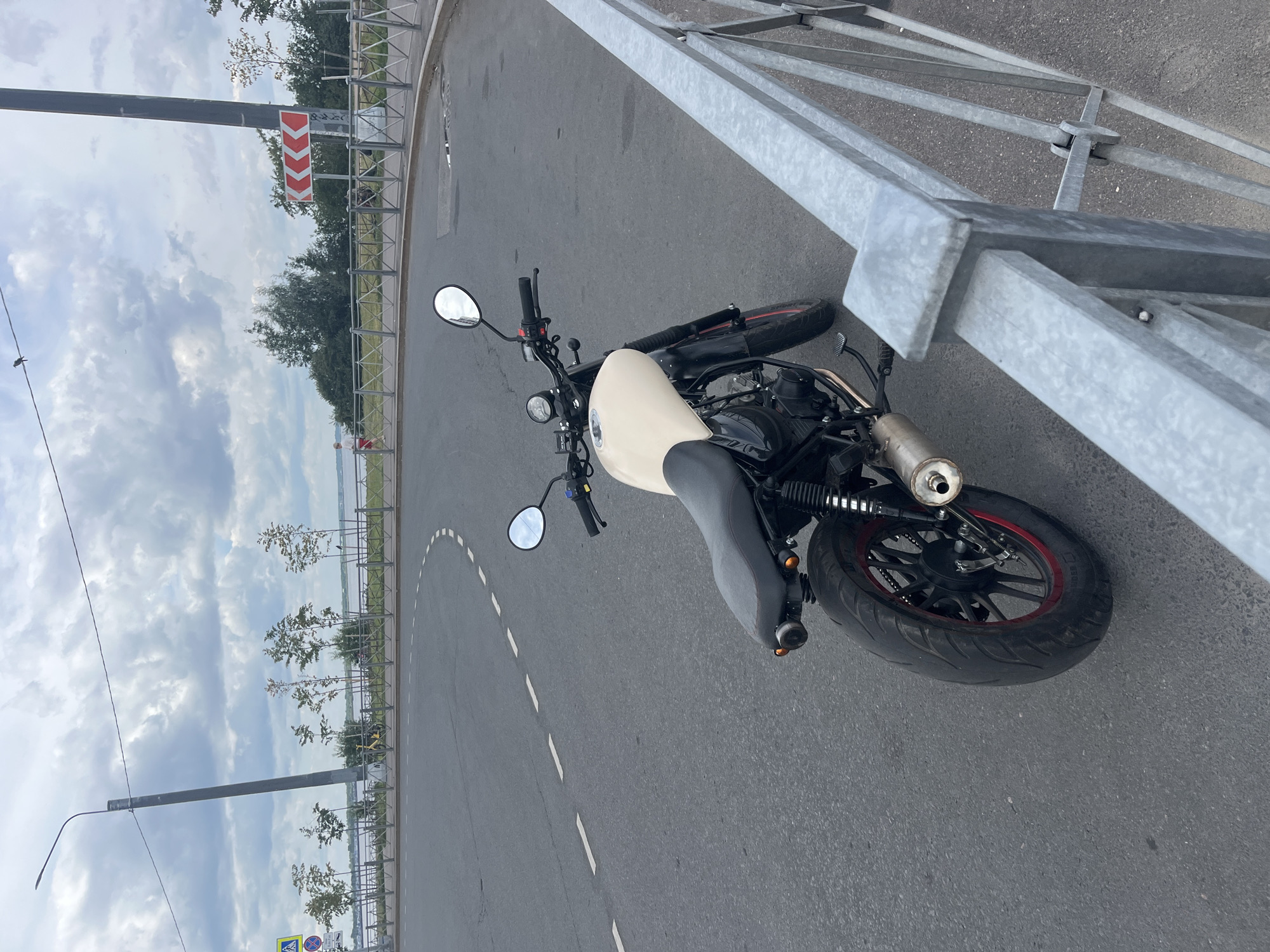 A cream-colored motorcycle is parked on a paved road with a white dashed line on the left. The motorcycle has a black seat, black handlebars with mirrors, and a chrome exhaust pipe. A grey metal fence is visible in the foreground to the right. In the background, there is a body of water with trees and a cloudy sky. A red and white directional sign with chevron arrows indicating a sharp left turn is mounted on a pole in the distance, along with a street light. A person in red clothing is walking near the water. The scene is likely in Saint Petersburg, Russia, near a waterfront area with a road and pedestrian access.