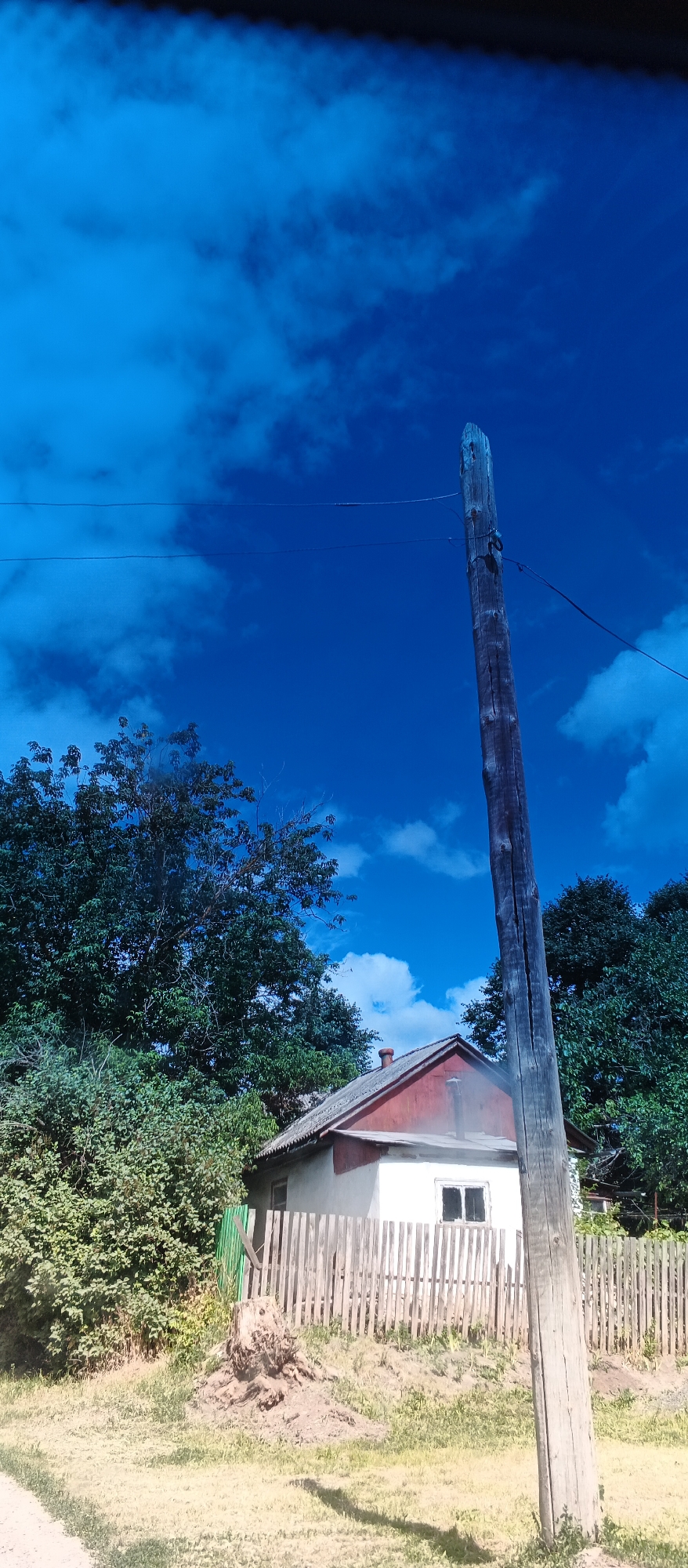 A vertical, rustic wooden utility pole stands prominently in the foreground, slightly off-center to the right, against a bright blue sky with scattered white clouds. Power lines are visible stretching horizontally across the upper portion of the frame, one seemingly attached to the pole. To the left and behind the pole, a cluster of mature green trees forms a dense backdrop. In the middle ground, a small, simple white house with a red-tiled roof and a red trim around the eaves is visible. A weathered wooden fence encloses the property in front of the house. A tree stump is situated to the left of the house, in front of the fence. The ground in front of the house and extending to the foreground is covered in dry grass and dirt. The overall scene depicts a rural or semi-rural setting.