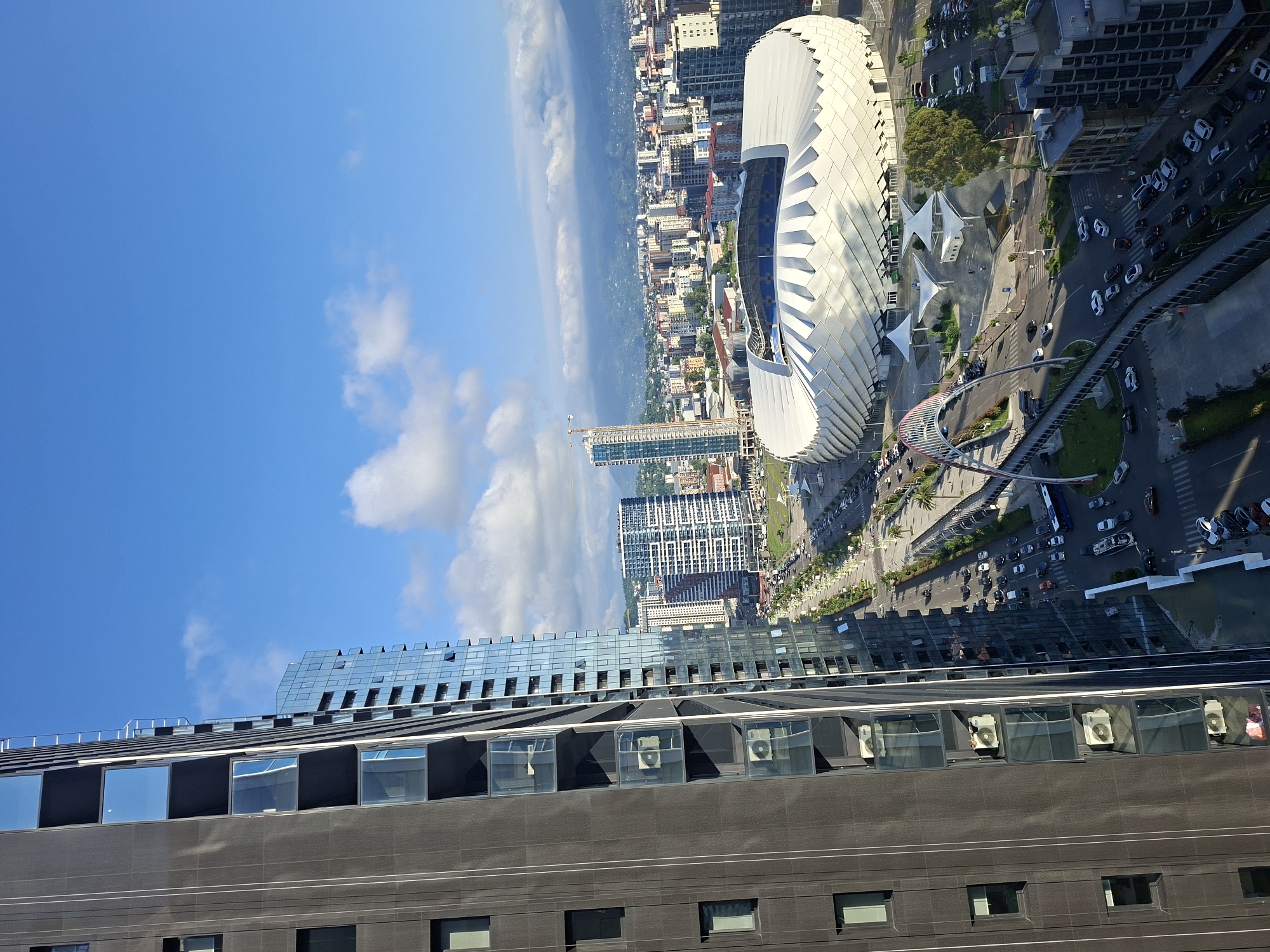 The image shows a city skyline on a clear day, with a focus on a modern stadium and surrounding urban development. In the foreground, a tall, dark-colored building with a grid of windows occupies the left side of the frame. To its right, a glass-fronted building reflects the sky and adjacent structures.
The central part of the image is dominated by a large, white, futuristic stadium with a distinctive patterned roof. An arching pedestrian bridge, red and white, spans a road in front of the stadium. A street with moderate traffic, including cars and a blue bus, runs alongside the stadium.
Beyond the stadium, a dense urban landscape of various buildings stretches towards the background, with mountains visible in the distance under a blue sky with scattered white clouds. The scene appears to be a vibrant, contemporary urban area.
Based on the architectural style of the stadium and surrounding buildings, this location is likely Batumi, Georgia, and the stadium is the Batumi Arena.
