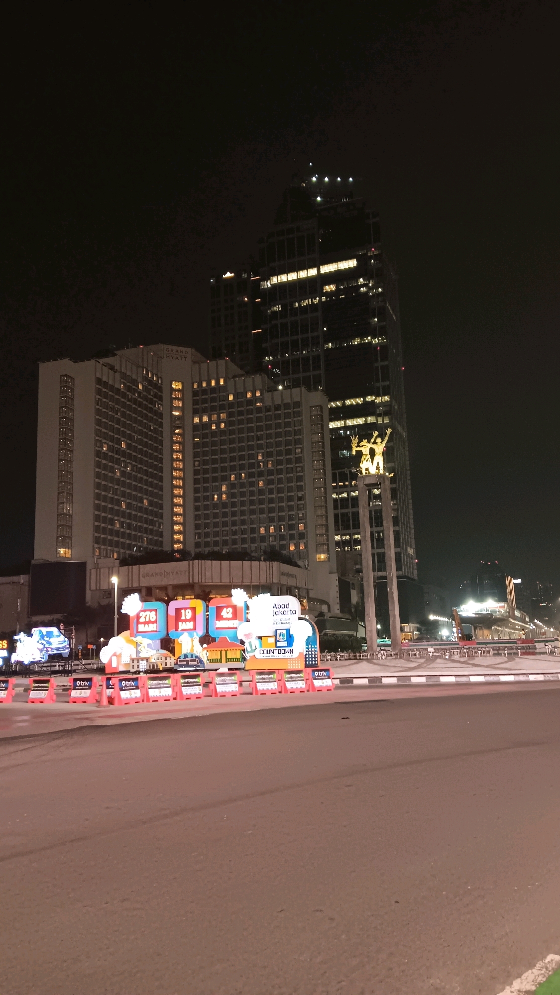 The image captures a night scene in Central Jakarta, Indonesia, specifically near the Hotel Indonesia Roundabout. In the background, the Grand Hyatt hotel stands tall, its windows illuminated, contrasting with the dark night sky. To its right, the iconic Arjuna Wiwaha statue, also known as the Hotel Indonesia roundabout statue, is visible, a landmark in the city. In the foreground, the street is lined with temporary barriers and decorative structures, possibly part of an event or countdown display, featuring colorful graphics and text like "Abad Jakarta" and "COUNTDOWN". The road itself appears to be asphalt. No people are clearly visible in the image. The overall impression is of a bustling urban environment at night, with prominent architectural structures and event decorations.
