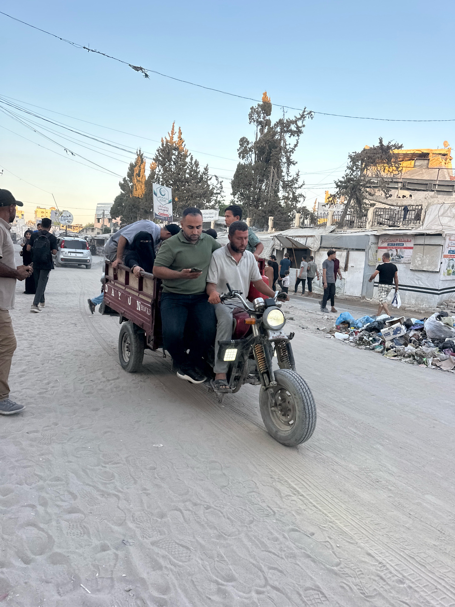 In the city of Gaza, Palestinian Territories, people are seen riding on a three-wheeled vehicle. The vehicle, which appears to be a cargo trike, is carrying several individuals, including a woman wearing a niqab and a man holding a mobile phone. The surrounding environment is characterized by a dusty, unpaved road, with buildings and tents visible in the background. Piles of garbage are scattered along the side of the road, and overhead power lines are present. The scene suggests a community navigating everyday life amidst challenging living conditions.