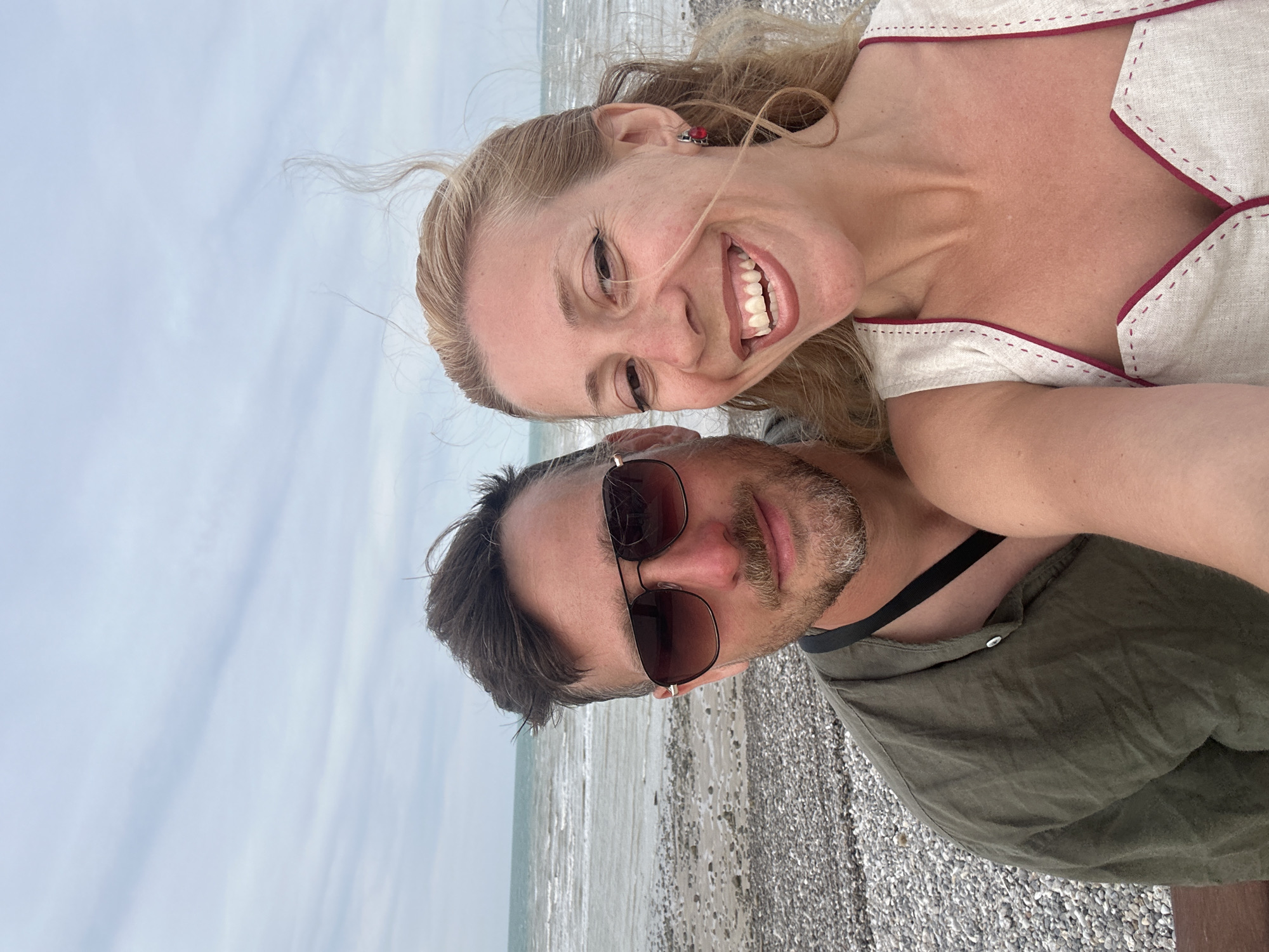 A couple takes a selfie on a pebble beach in Saint-Valery-en-Caux, France. The man, on the left, wears sunglasses and an olive-green shirt. The woman, on the right, smiles widely, her blonde hair blowing in the wind. She wears a cream-colored top with red trim. The sea and sky are visible in the background.