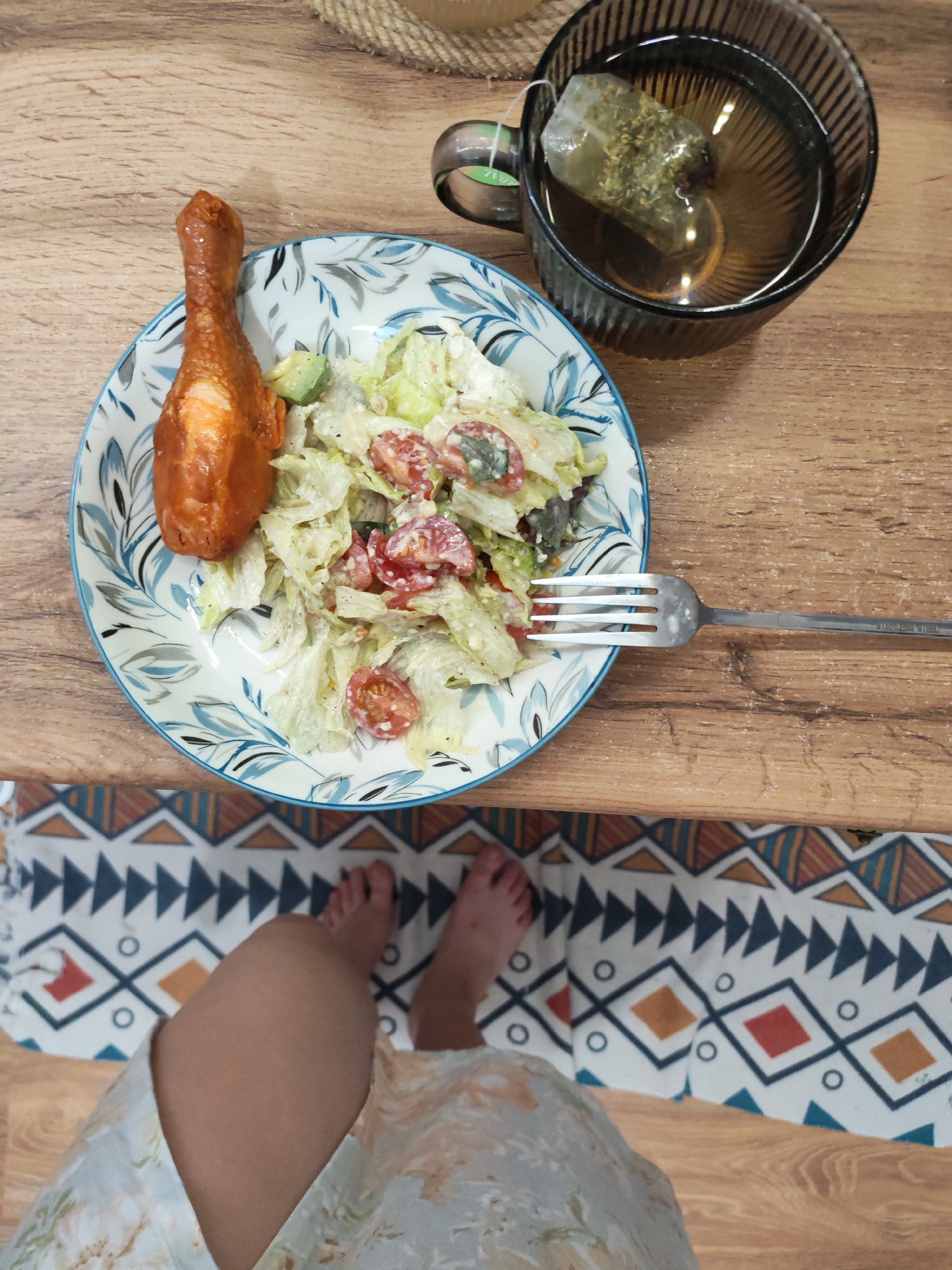 A close-up, overhead shot shows a meal on a wooden table. The meal consists of a chicken leg and a salad on a plate with a blue and white floral pattern. A fork rests on the edge of the plate. To the right of the plate is a clear glass mug filled with tea and a tea bag. The background shows a woven placemat and a patterned rug. The image is taken from a low angle, showing a person's legs and feet wearing a floral dress. The scene is indoors, on a wooden table, with a colorful rug on the floor. The lighting suggests it is daytime.