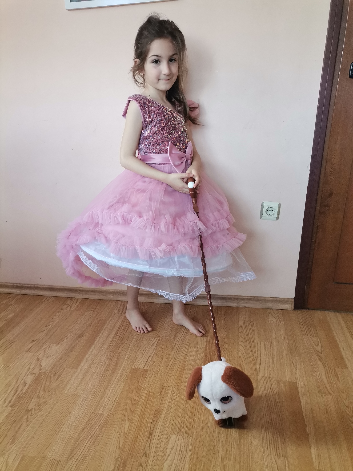 A young girl stands in a room with wooden floors and light pink walls, wearing a pink sequined dress with a ruffled skirt and a matching pink sash. She holds a brown walking stick with a white knob at the top. Attached to the bottom of the walking stick is a plush toy dog with white fur and brown ears. The girl is barefoot. There is a white electrical outlet on the wall behind her, and a dark wooden door is visible on the right side of the frame. A white-framed picture hangs above her on the wall. The location is Unknown, Unknown.
