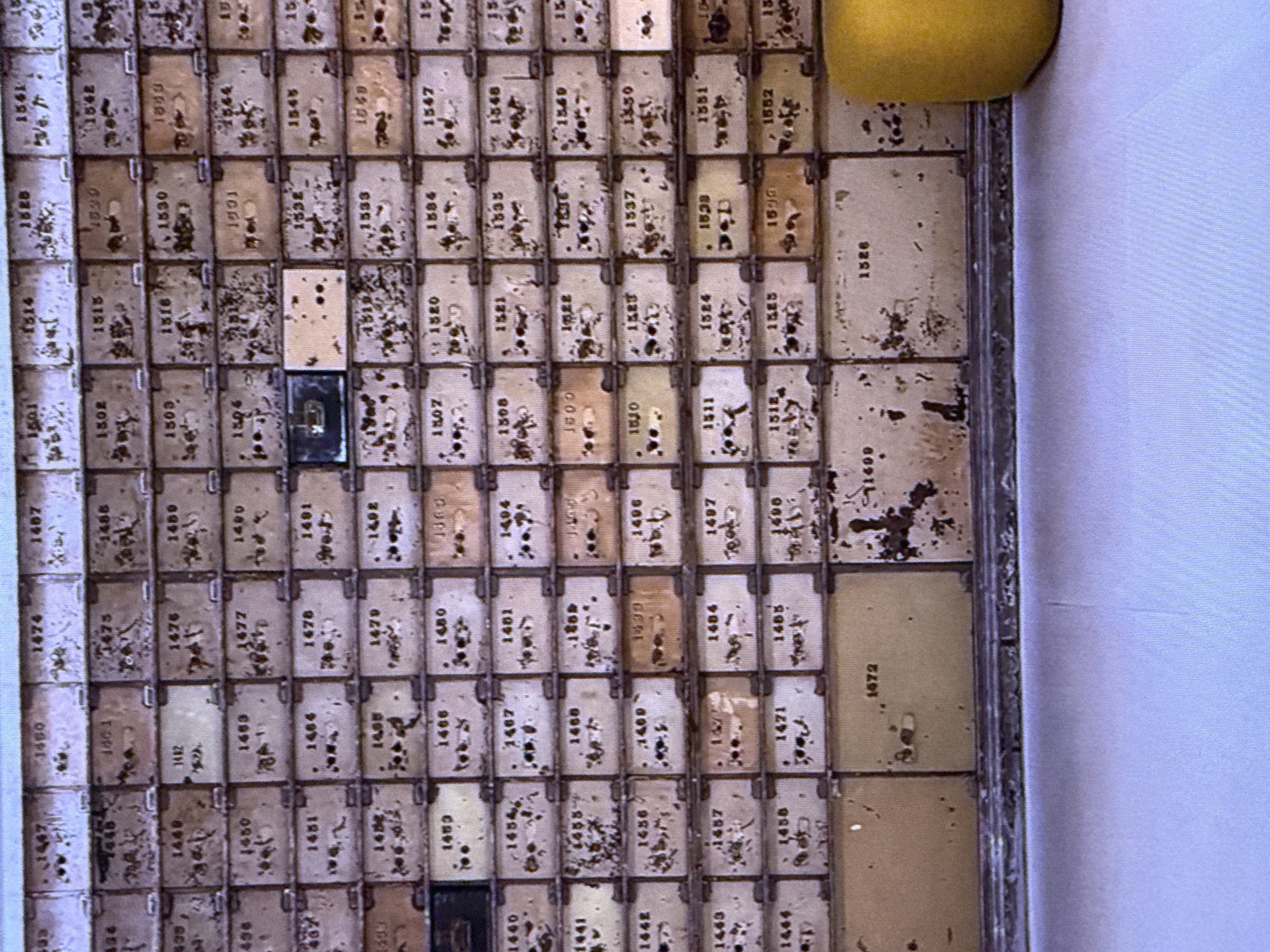 The image displays a section of a wall with a grid of small, numbered compartments. The compartments are worn and show signs of age, with some appearing to have rust or paint damage. Each compartment has a number printed on it, ranging from the 1400s to the 1500s. In the lower right corner of the image, a bright yellow object, possibly a piece of fruit, is partially visible. The background is out of focus and appears to be a light-colored surface. The scene is not indicative of any specific location in Dubai, United Arab Emirates, as it lacks distinguishing architectural features or landmarks.