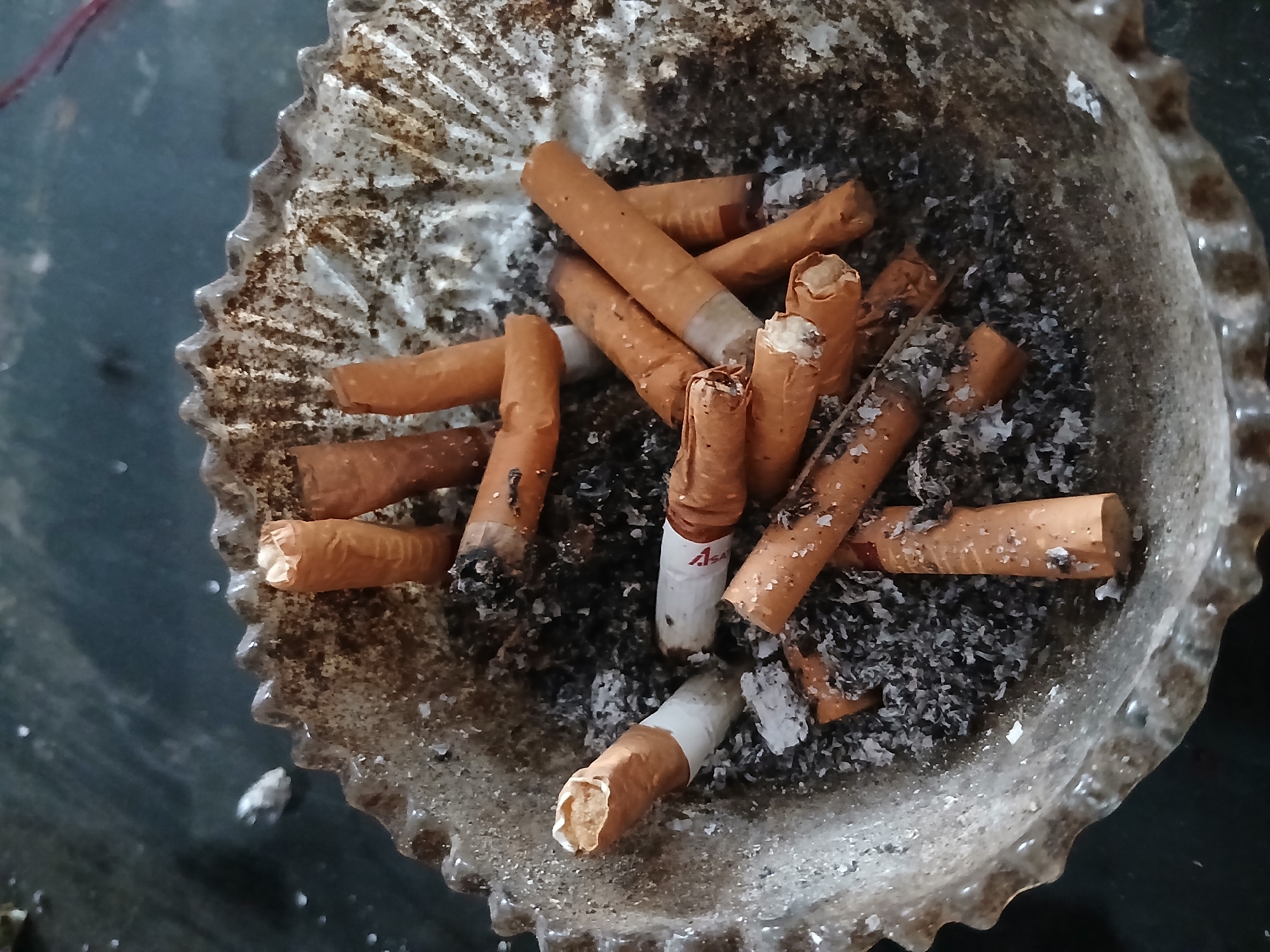 In Kademangan Kulon, Indonesia, an ashtray filled with cigarette butts and ash sits on a dark, textured surface. The ashtray itself is made of clear, wavy glass and appears to be quite full. The cigarette butts are of various stages of being smoked, with some appearing freshly extinguished and others more heavily used, their filters exposed. The ash is a mixture of gray and black, scattered throughout the ashtray and around the butts. The lighting is dim, casting subtle shadows across the scene.