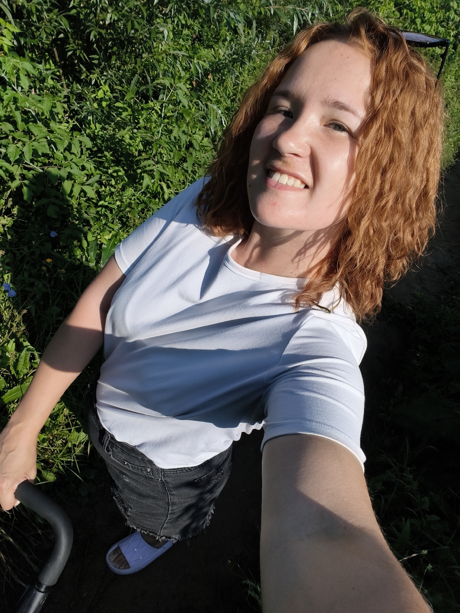 A young woman with curly auburn hair, wearing a white t-shirt and black denim shorts, is taking a selfie outdoors. She is smiling, and her sunglasses are perched on top of her head. The background consists of lush green foliage, suggesting a natural or park-like setting. The lighting indicates it is daytime, with harsh shadows cast by the sun. The woman is holding onto a black handle, possibly from a stroller or a similar object. She is wearing blue slides on her feet. The overall scene is casual and appears to be taken in an outdoor, potentially grassy or wooded area.