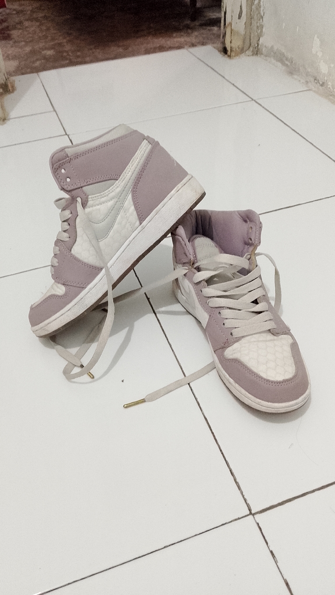 Two high-top sneakers, one on top of the other, are positioned on a tiled floor. The sneakers are predominantly light purple and white, with white laces. The shoe on the bottom is facing forward, while the shoe on top is angled with its toe pointing upward. The tiled floor appears to be white with thin gray grout lines. In the background, a wall with peeling paint and a darker area that could be furniture or flooring is visible. The scene is indoors, likely a room. The location specified, Seputih, Indonesia, is not discernable from the visual information present in the image.