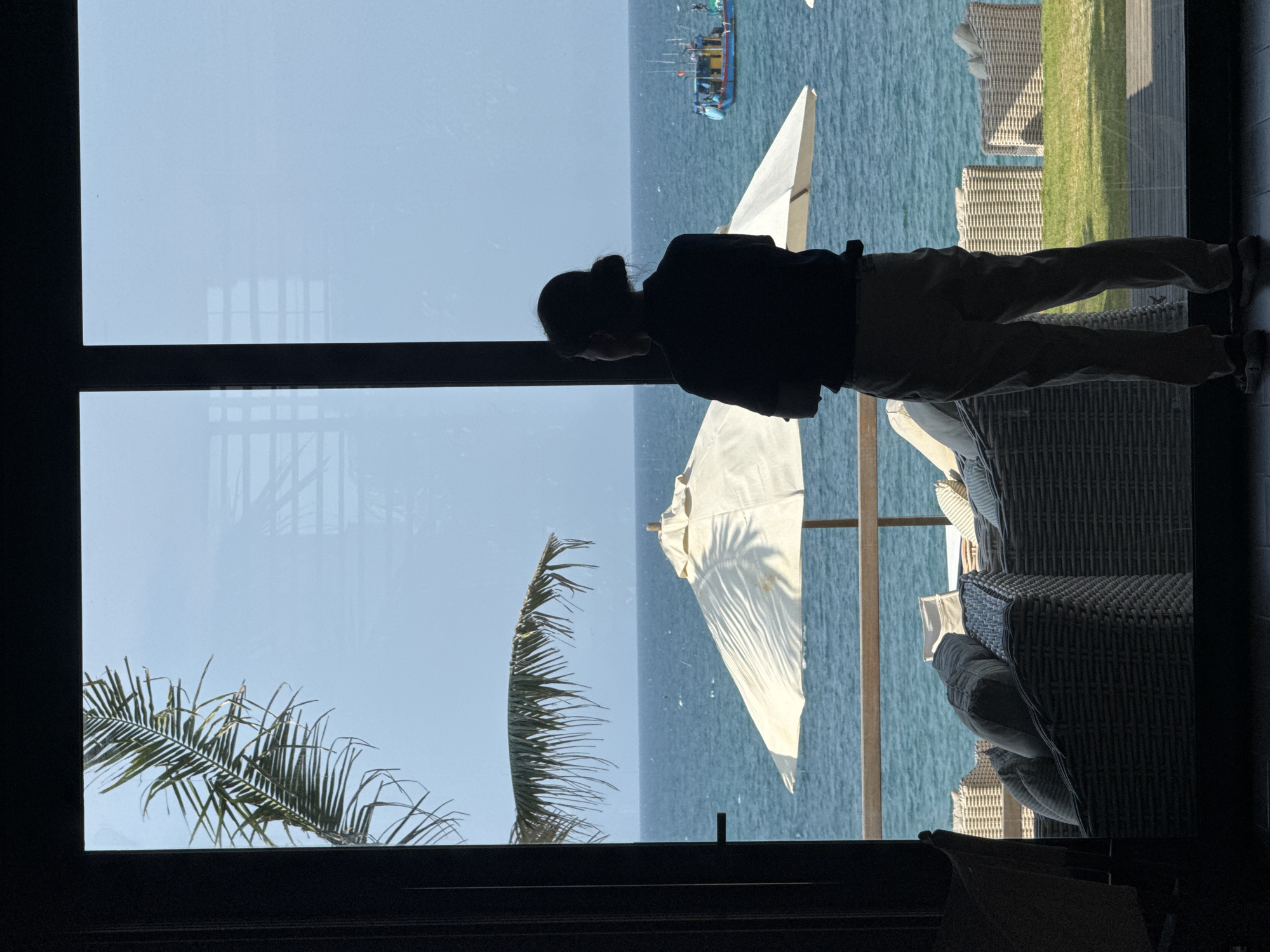 A person stands facing an ocean view visible through a large window. The person has dark hair pulled back and is wearing dark pants and a dark shirt. Outside the window, a palm frond is visible on the left, and white beach umbrellas and chairs are present on the right. The ocean appears blue and calm, with a small boat visible in the distance. Đà Nẵng, Vietnam cannot be confirmed as the location from the image alone.