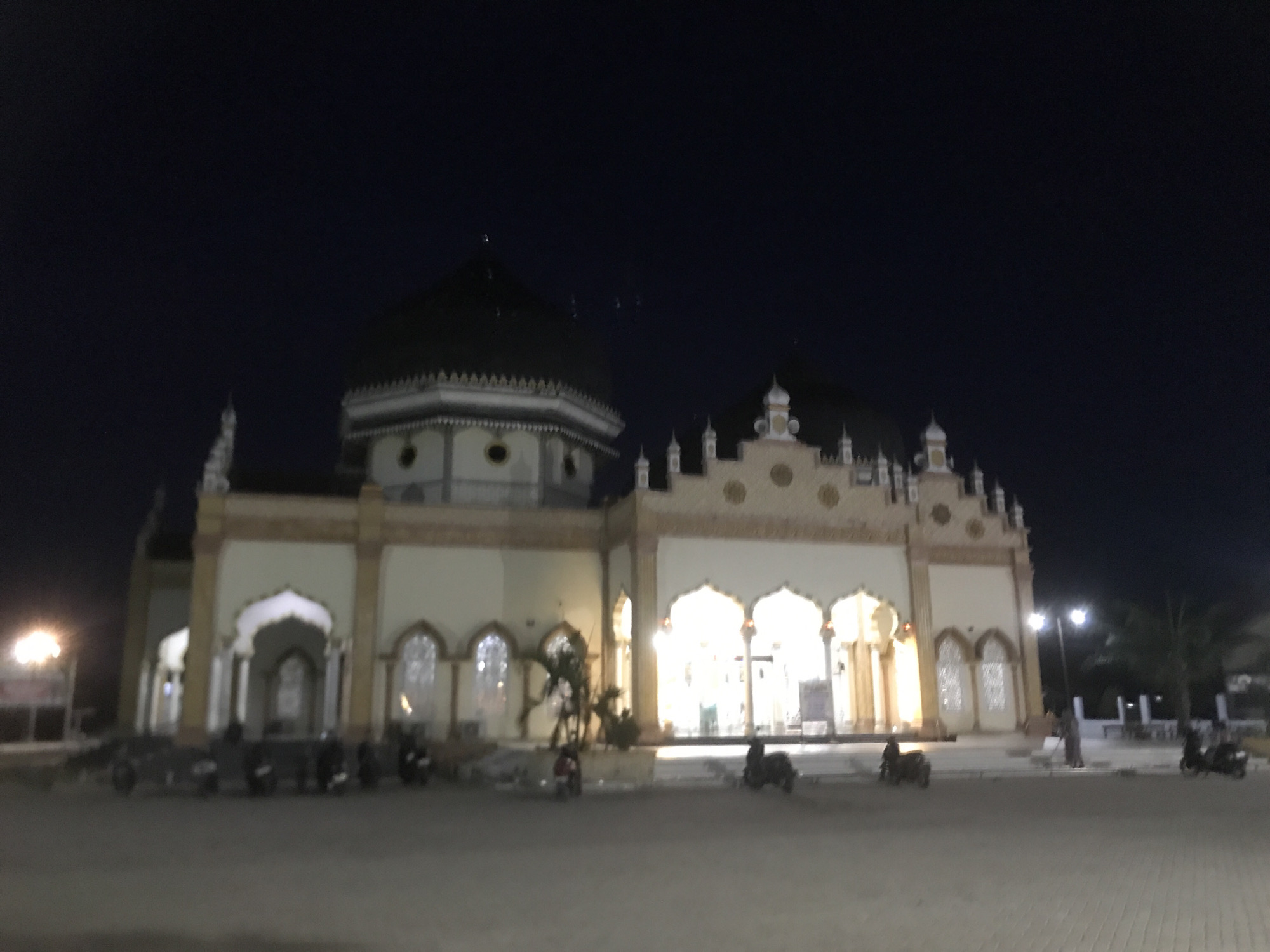 At night, the Great Mosque of Keude Peureulak is illuminated, its cream-colored facade and dark dome contrasting against the night sky. Motorbikes are parked in the foreground. A figure sweeps the paved area in front of the mosque. Streetlights illuminate the scene.