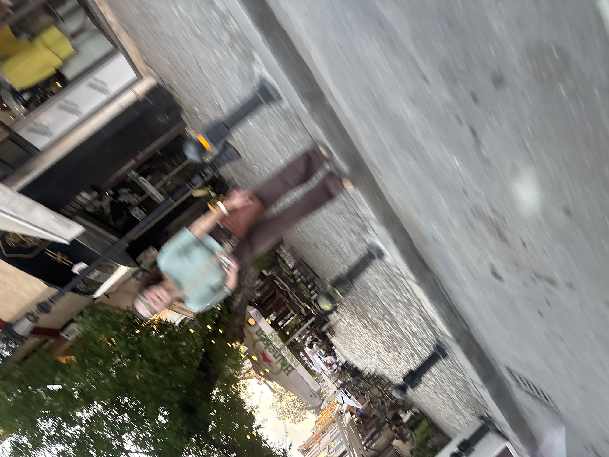 This blurry and rotated image, taken in what is specified as Plovdiv, Bulgaria, captures an outdoor urban scene, likely an evening at a cafe or restaurant. The primary subject is a person, possibly female, positioned in the middle-left of the frame. They are wearing a light blue or teal top and brown pants, with their face partially obscured by blur. They appear to be holding a white object, possibly a cigarette, in their right hand.
The setting includes an outdoor seating area with visible, albeit blurred, tables and chairs. A large white umbrella prominently features the green "Heineken" text and its red star logo. Several dark-colored lampposts with glowing yellow lights are scattered throughout the scene, indicating it's likely dusk or evening. Lush green foliage, probably trees or bushes, with small, warm-toned fairy lights, is visible in the lower left, adding to the ambiance. Parts of buildings or storefronts line the left side and background, with some windows reflecting light or showing interior details. The ground is a light-colored paved surface, consistent with a street or pedestrian area. A small portion of a white vehicle is visible in the bottom right corner. The image quality is heavily blurred and rotated 90 degrees counter-clockwise, impacting clarity.