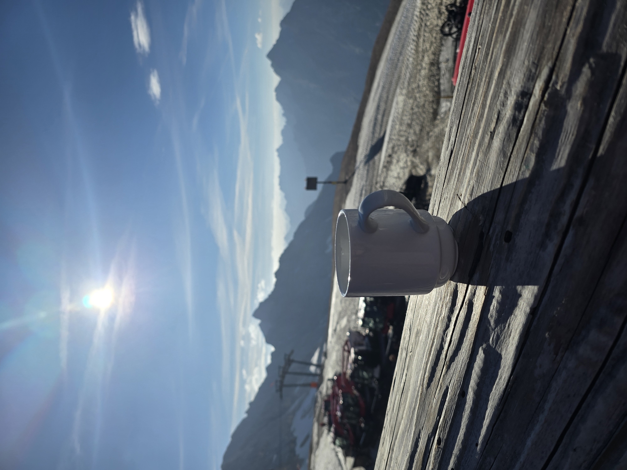 The image shows a white mug on a weathered wooden surface in the foreground. In the background, a panoramic view of mountainous terrain is visible under a bright, sunny sky with some wispy clouds. The sun is prominent in the upper left quadrant of the sky, casting lens flares. Snow is present on the slopes, and several red snow groomers are parked on the terrain. Ski lift pylons are also visible in the mid-ground. The scene appears to be a scenic overlook or a rest stop at a mountain resort, possibly in the Bavarian Alps near Garmisch-Partenkirchen, Germany, given the mountainous landscape and the presence of ski equipment. There are no people visible in the image. The overall impression is one of a peaceful, elevated vantage point.