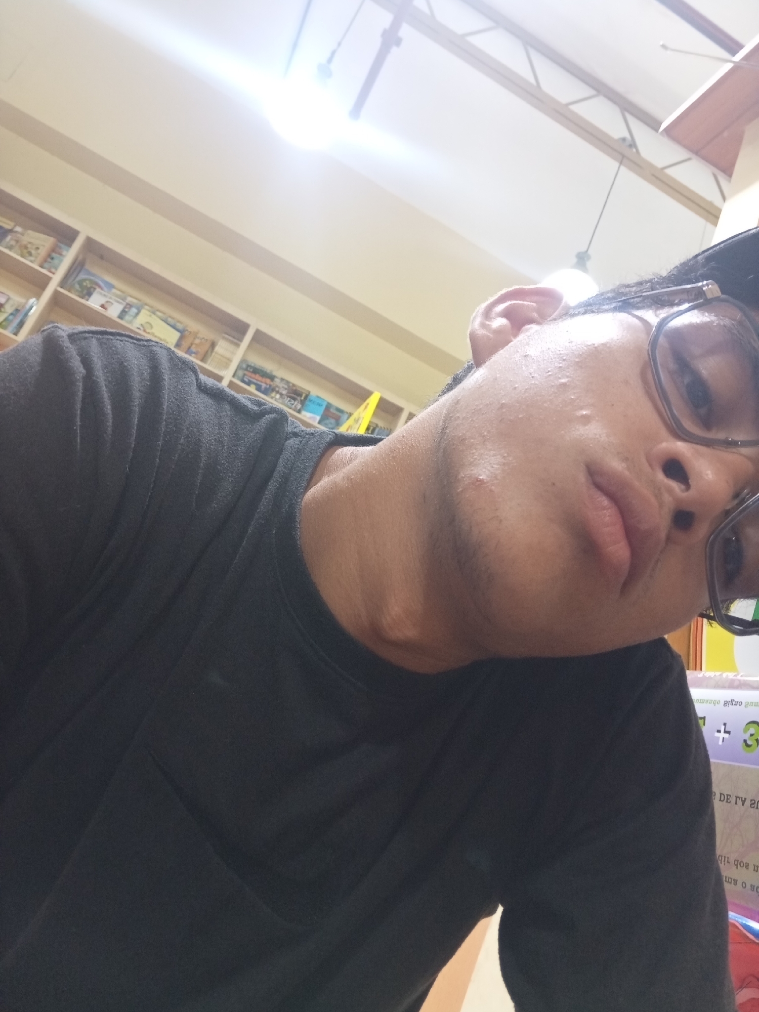 In Juan Griego, Venezuela, a young person is captured in a close-up, slightly tilted selfie. They are wearing a dark t-shirt and eyeglasses. The background reveals bookshelves filled with books, suggesting a library or bookstore setting. The lighting is bright, with a light fixture visible overhead. The focus is on the person's face, with their skin texture and some blemishes clearly visible. The overall impression is a casual, personal snapshot taken in an indoor environment.
