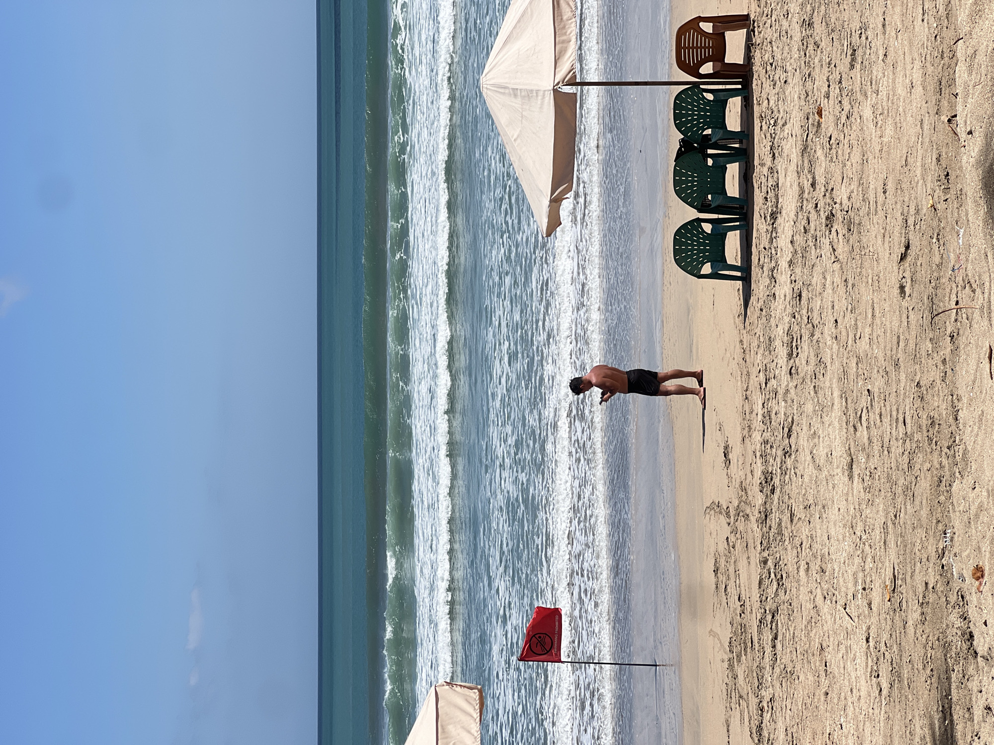A man stands on a sandy beach in Kuta, Indonesia, holding a mobile phone. He is wearing black swim trunks. In front of him, a red flag with a white symbol and the words "SWIMMING PROHIBITED" is planted in the sand. To the right, a beige beach umbrella shades four plastic chairs: three green and one brown. The ocean, with its characteristic turquoise water and white-capped waves, stretches out to the horizon under a clear blue sky. The beach itself is sandy with some debris scattered across the surface.
